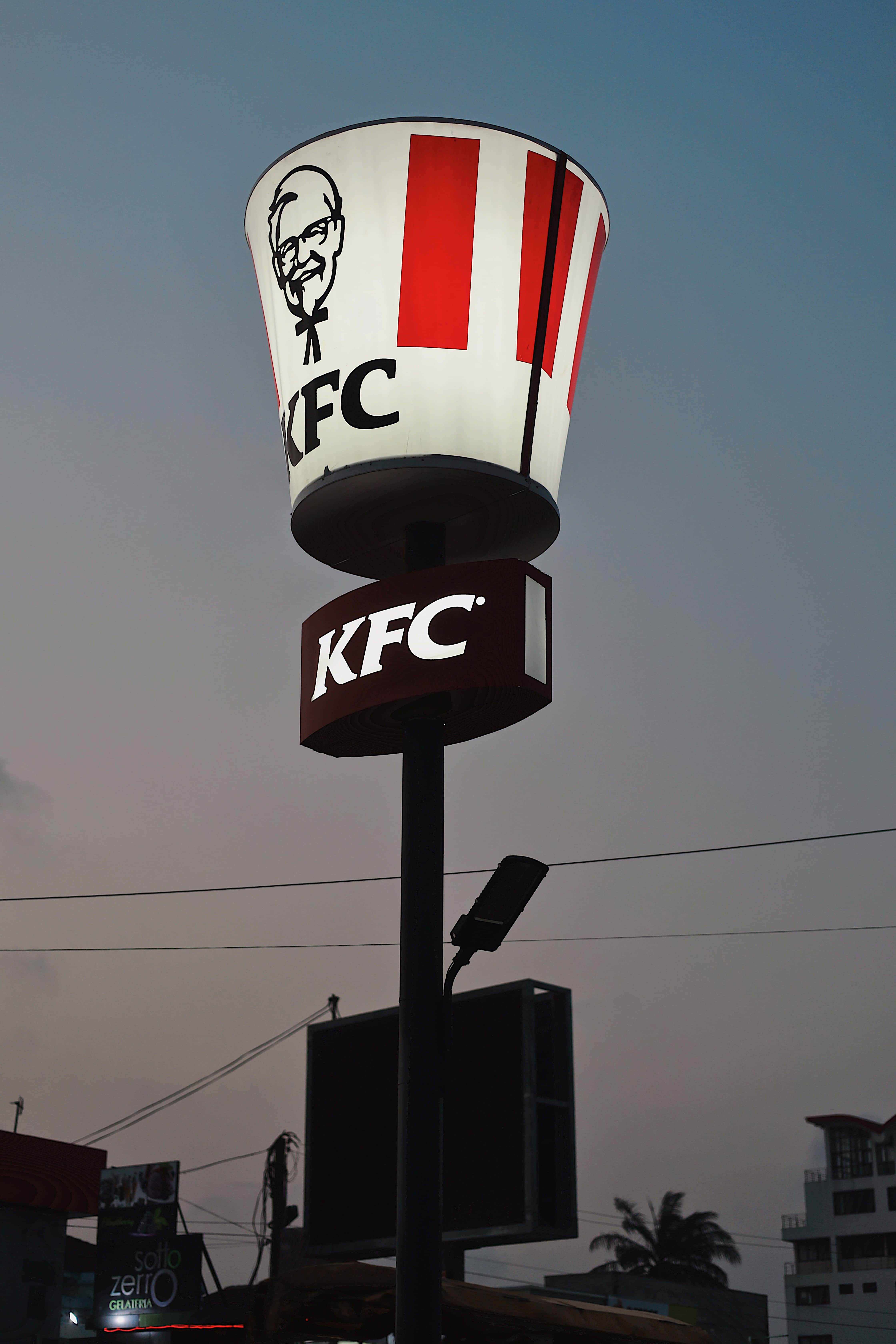 城中最美味的炸雞 壁紙 — 一個發光的KFC桶形招牌聳立在黃昏的天空下，背景有城市建築物和棕櫚樹。