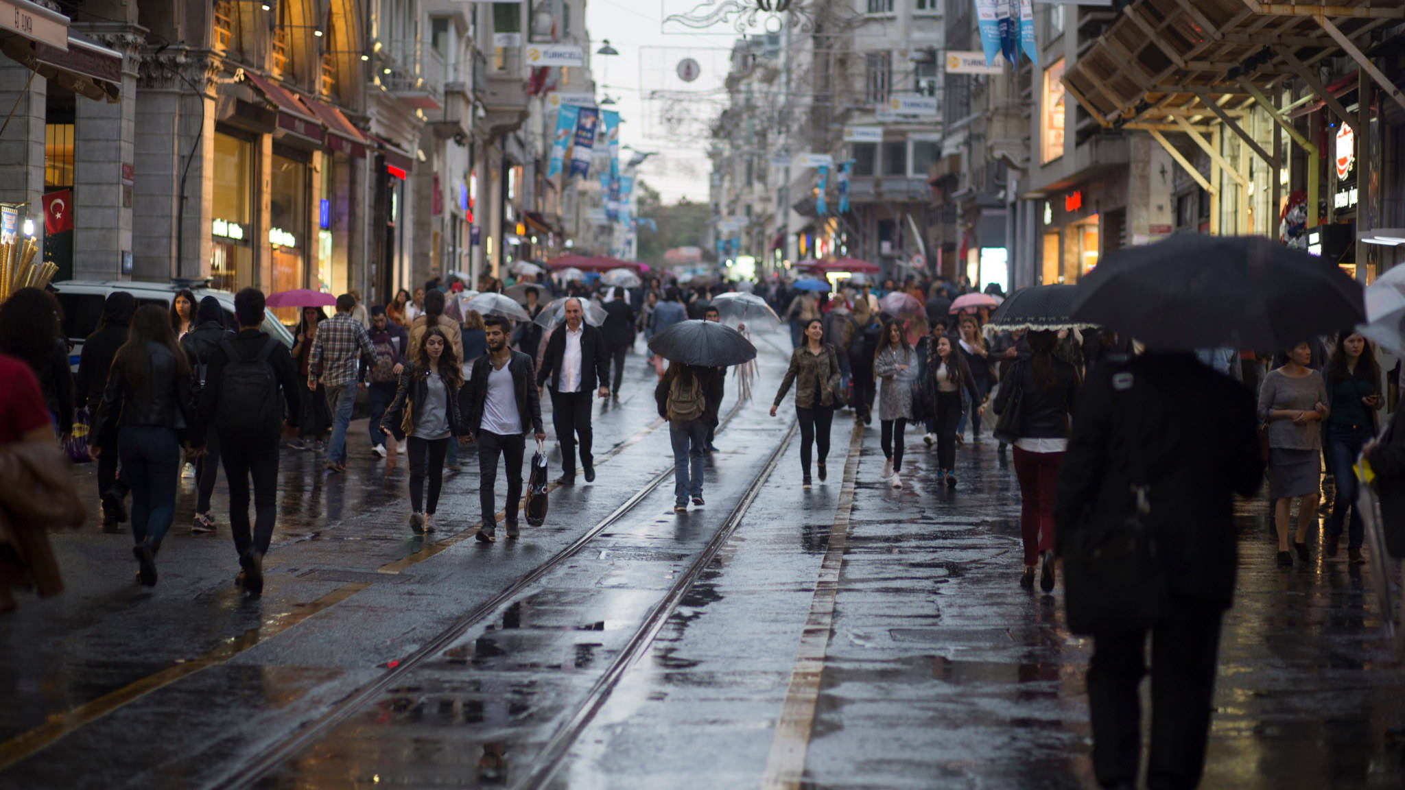城市中的人群 壁紙 — 行人撐著雨傘，在陰天之下，沿著兩旁商店和電車軌道的濕滑城市街道前行。