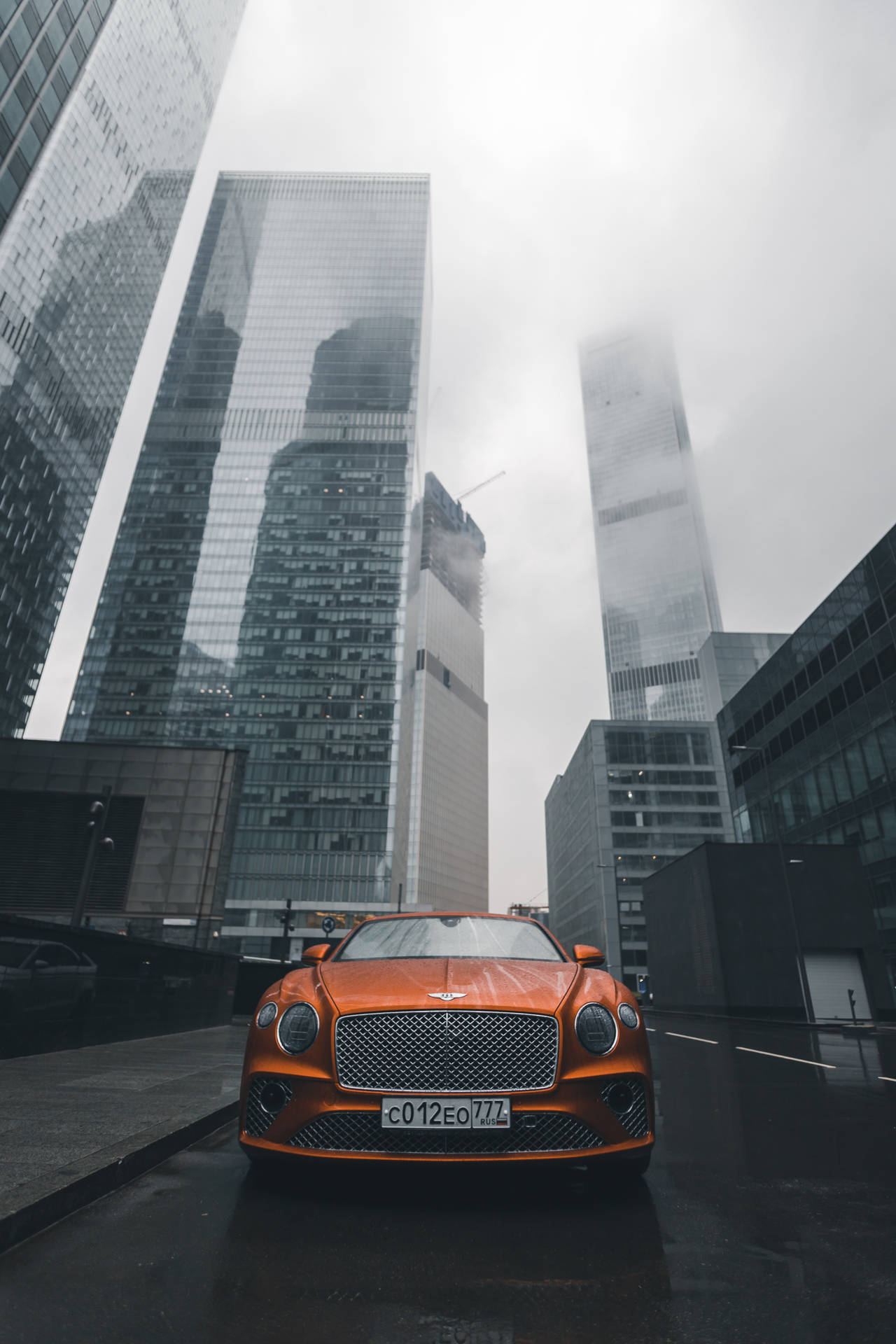 橙色豪華汽車停泊在濕潤的城市街道上，兩旁是高樓玻璃建築。