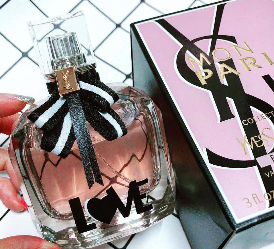 城市中的高級時尚 — YSL 壁紙 — 一隻手拿著帶有黑白條紋蝴蝶結的YSL Mon Paris香水瓶，背景為幾何白色。