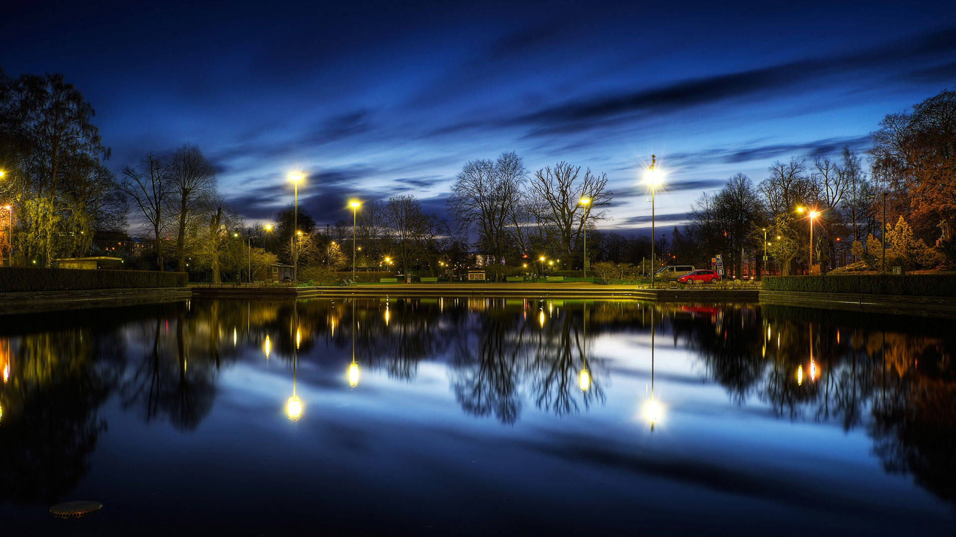 城市公園的美麗夜景 壁紙 — 一個寧靜的黃昏公園池塘，在深藍色的天空下映照著發光的街燈和光禿的樹木。