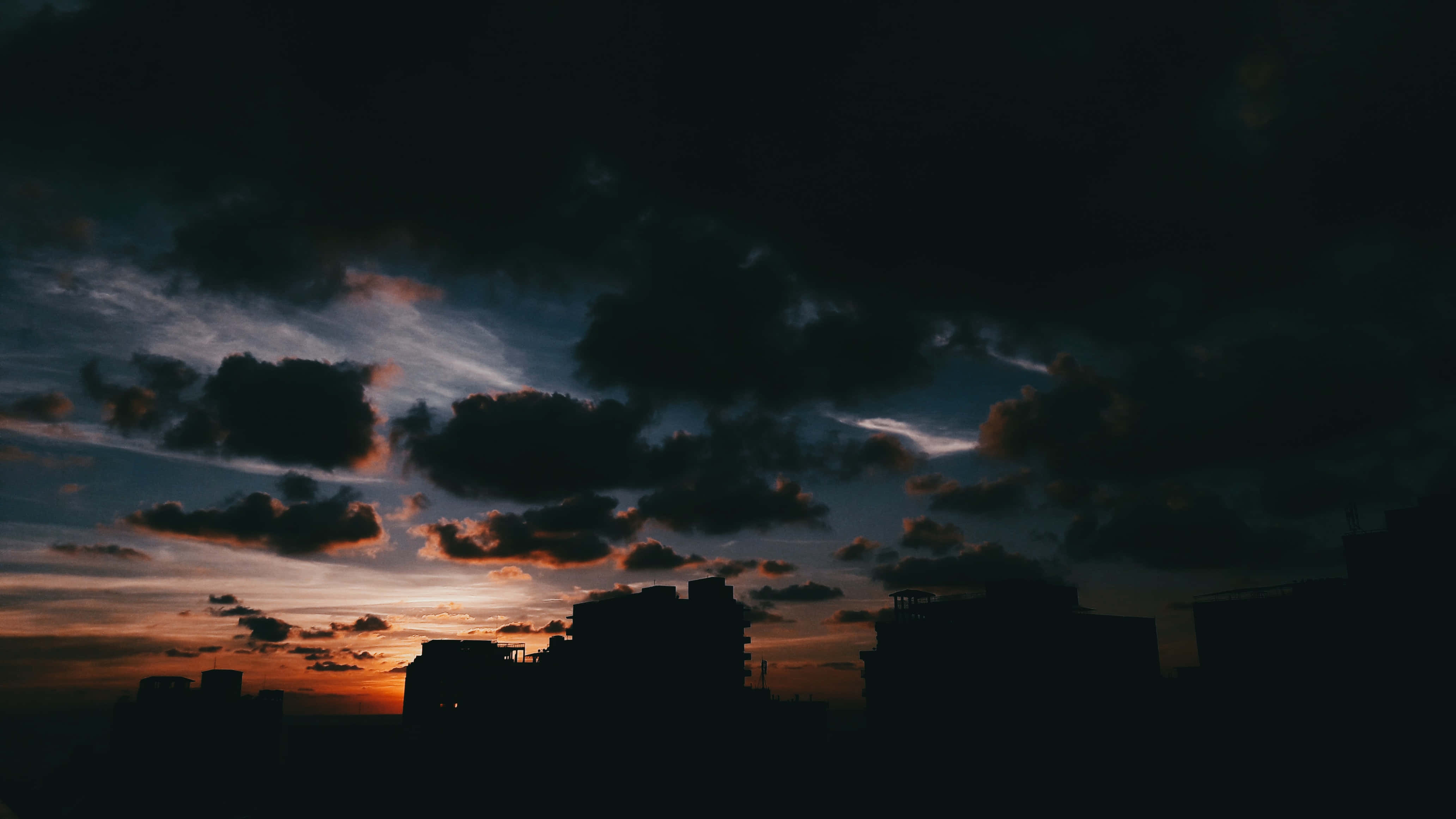城市剪影與烏雲 壁紙 — 城市天際線在戲劇性的暮色天空下形成剪影，天空中有火紅的橙色雲朵和深藍色的陰影。