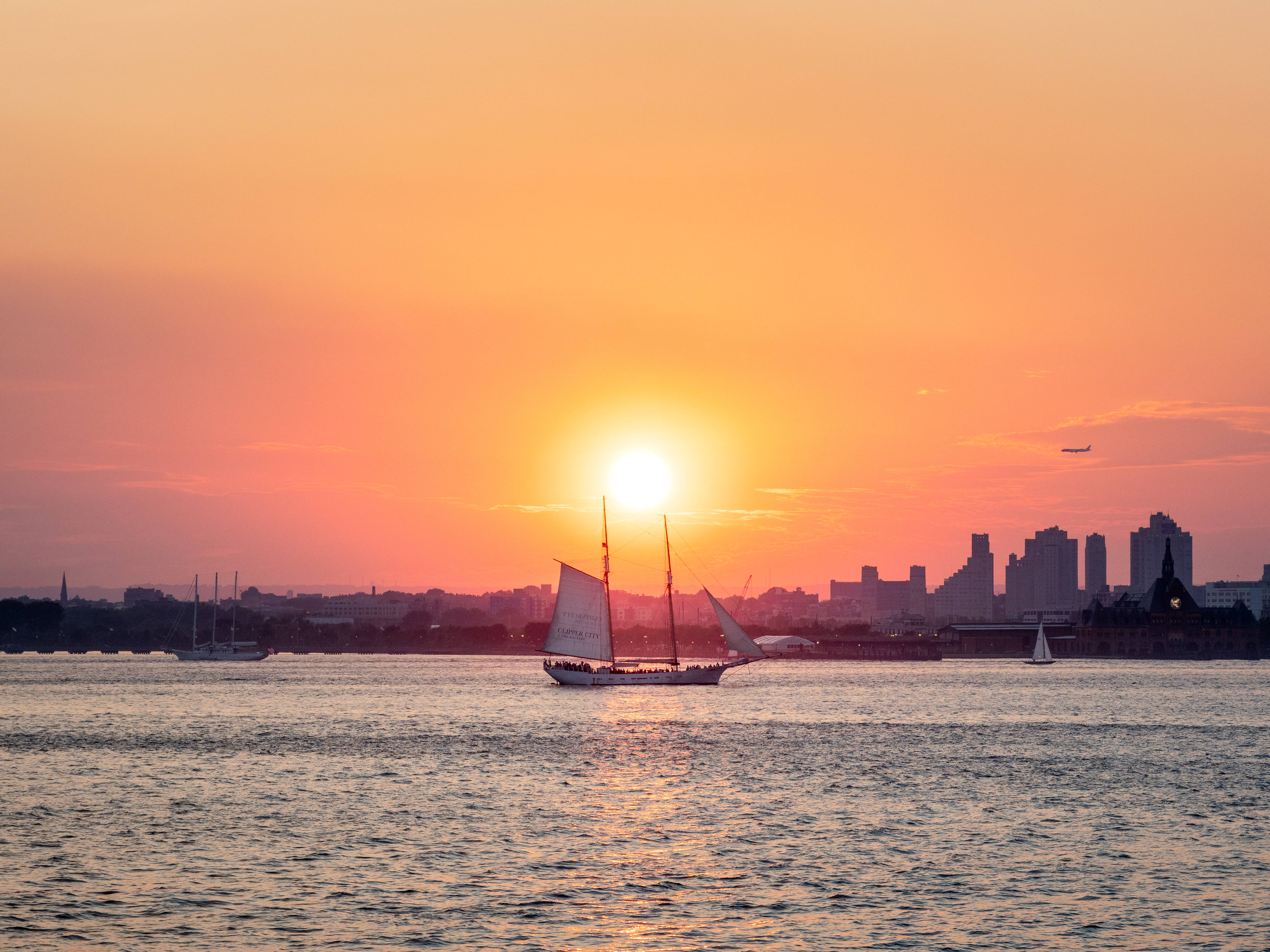 城市夕陽下的航行 壁紙 — 一艘帆船在日落時分滑過寧靜的港口，天空泛著橙色光芒，城市天際線呈現剪影。