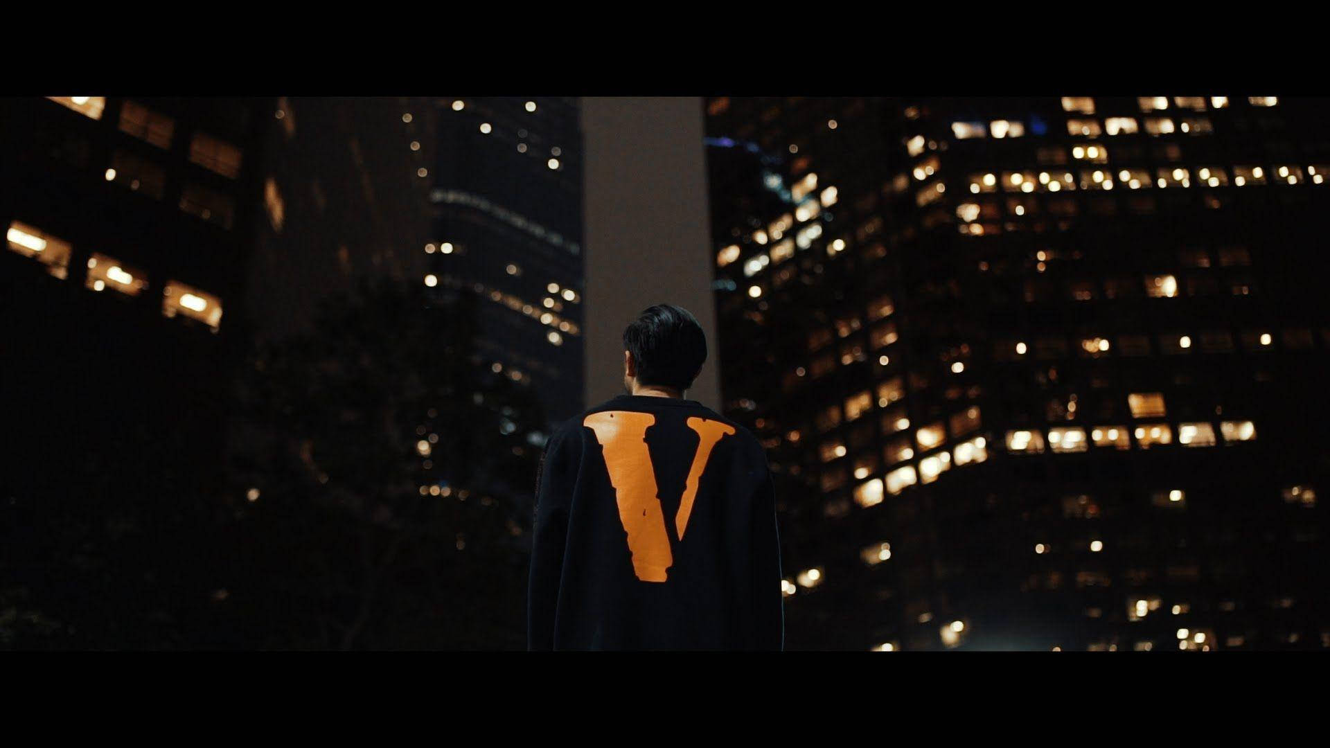 城市夜景中的橙色 VLONE 標誌 壁紙 — 一個身穿黑色斗篷、帶有發光橙色「V」的人獨自站在夜晚，凝視著燈火通明的摩天大樓城市景觀。