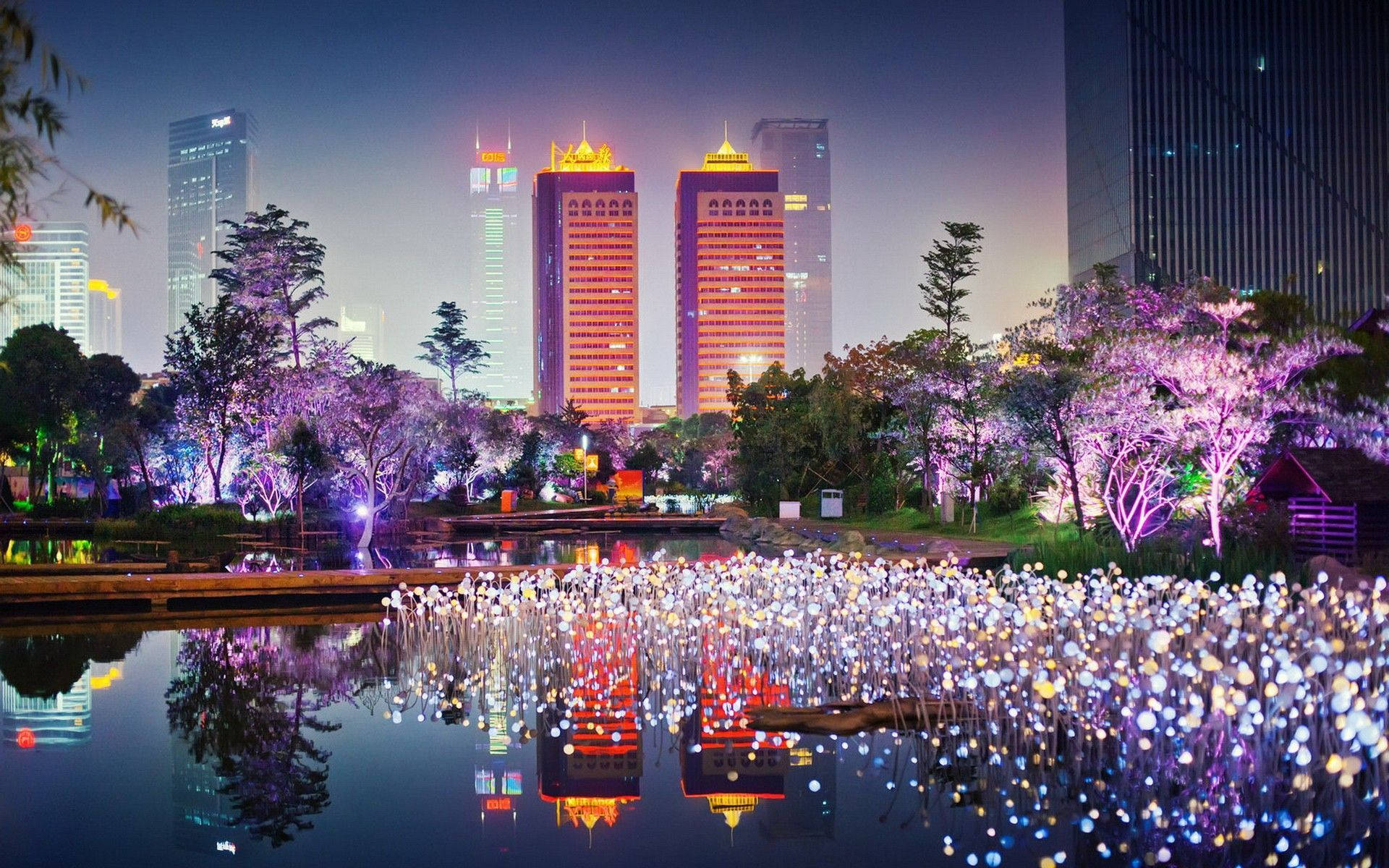 夜間城市公園，有發光的樹木和摩天大樓在水中的倒影。