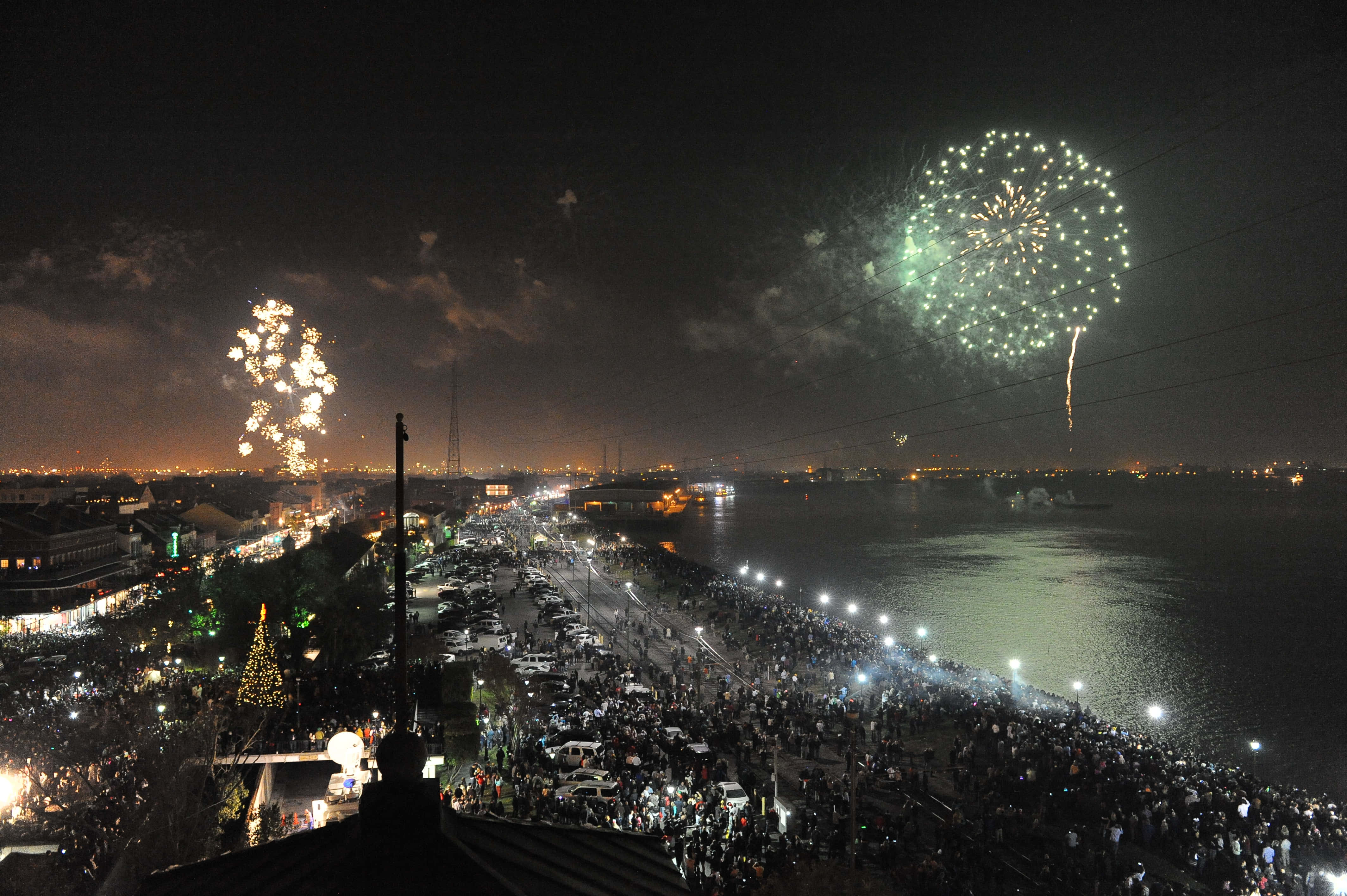 城市夜景新年圖 壁紙 — 在擁擠的海濱上空上演的充滿活力的夜間煙火表演，發光的爆炸反射在水面和下方的城市燈光上。