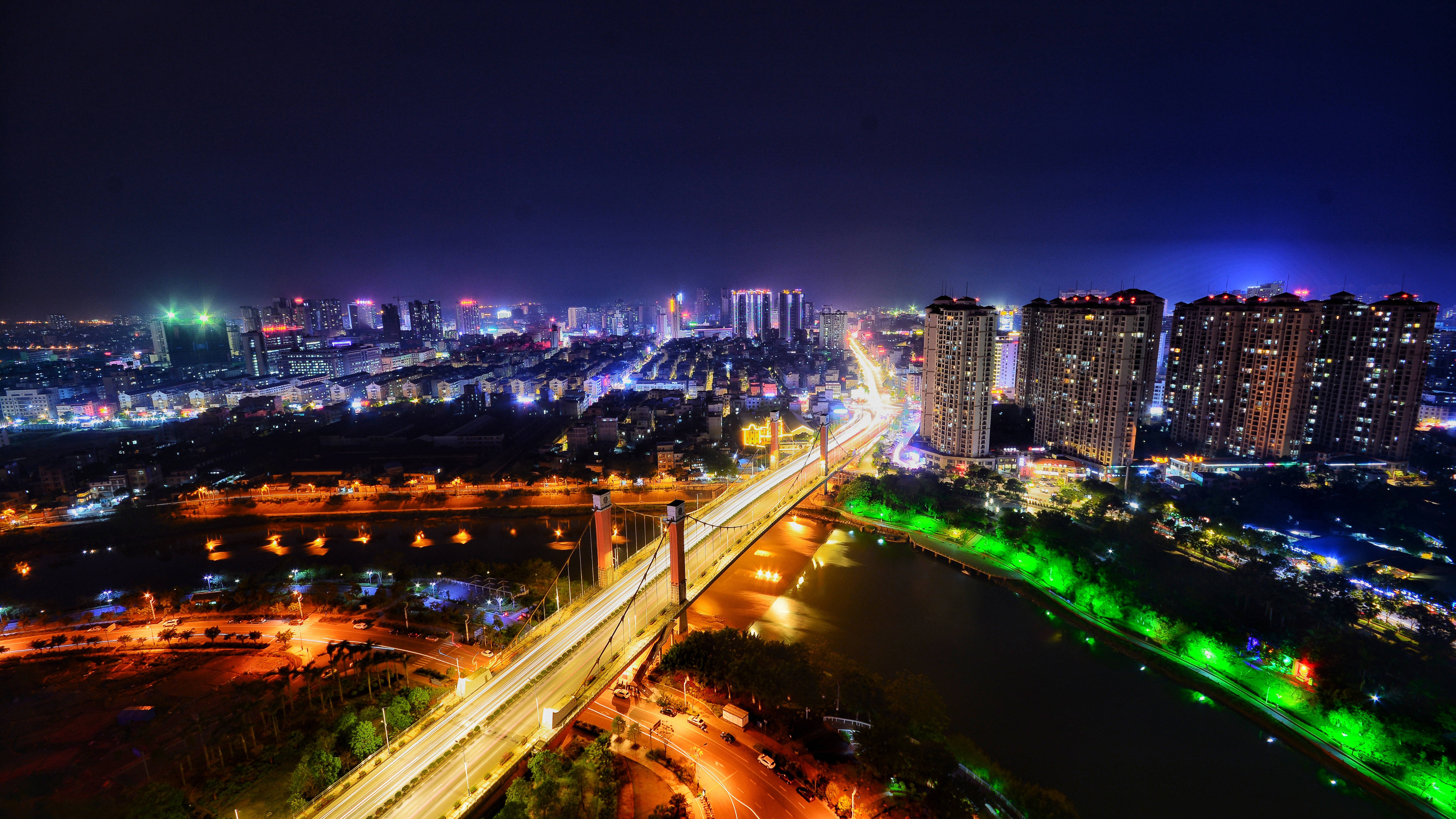 城市夜景燈光 4K 壁紙 — 充滿活力的城市景觀的夜間鳥瞰圖，有發光的摩天大樓、一座橫跨河流的燈光橋樑和車流的燈光軌跡。