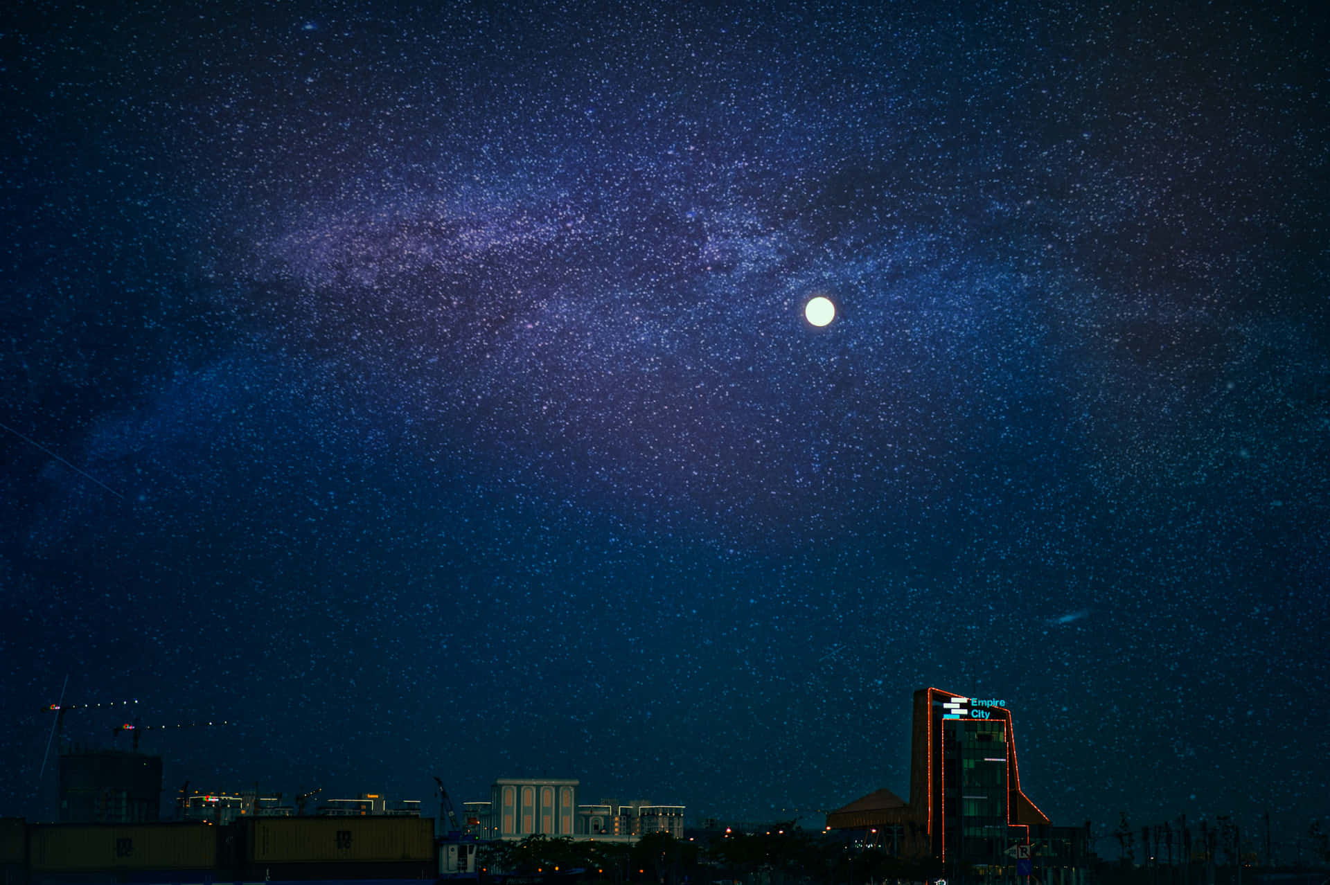 城市夜空中的月亮圖 壁紙 — 夜晚的城市天際線，下方是充滿活力的星空，明亮的月亮和銀河拱起。