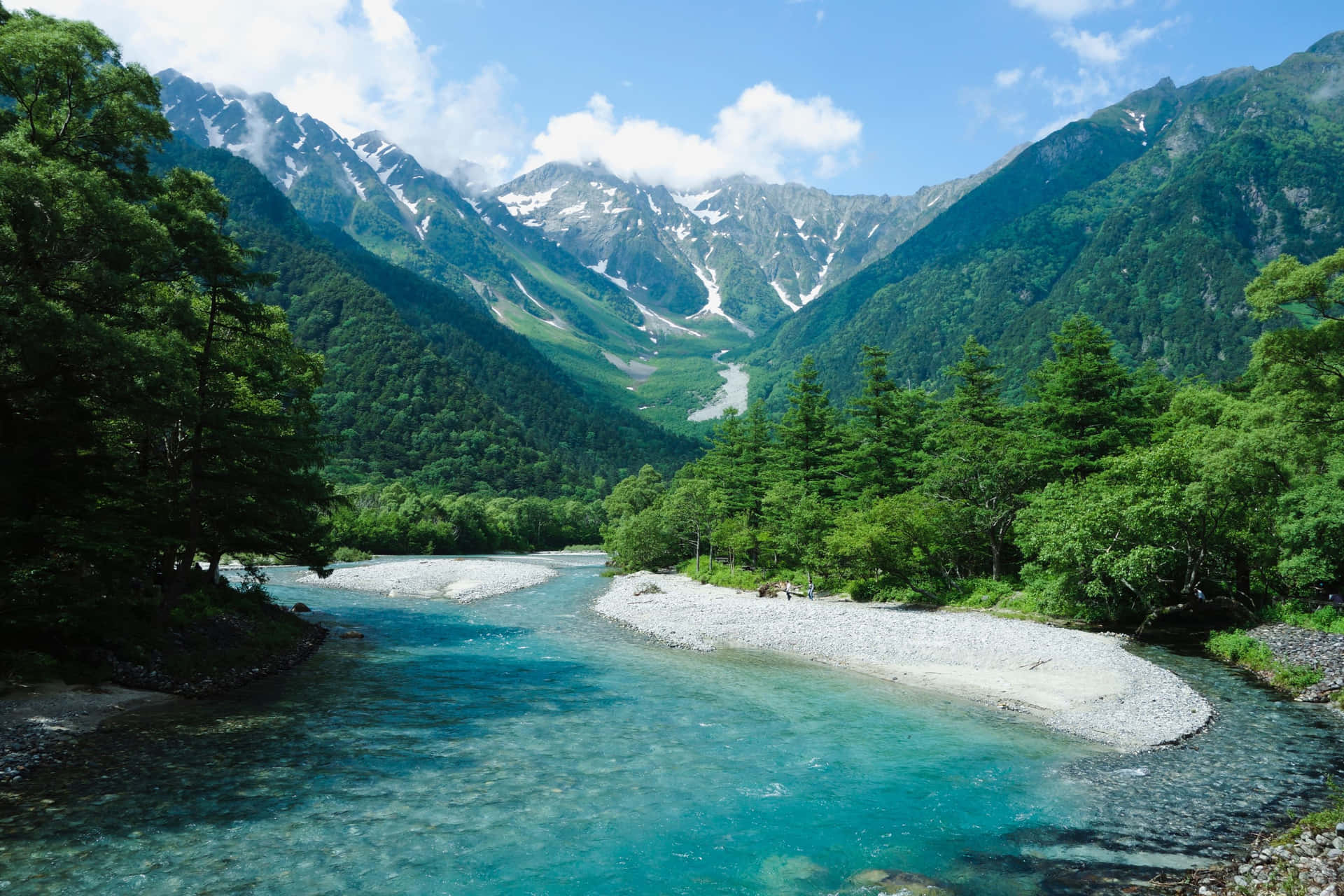 一條清澈的藍綠色河流流經森林覆蓋的山谷，背景是雪山。