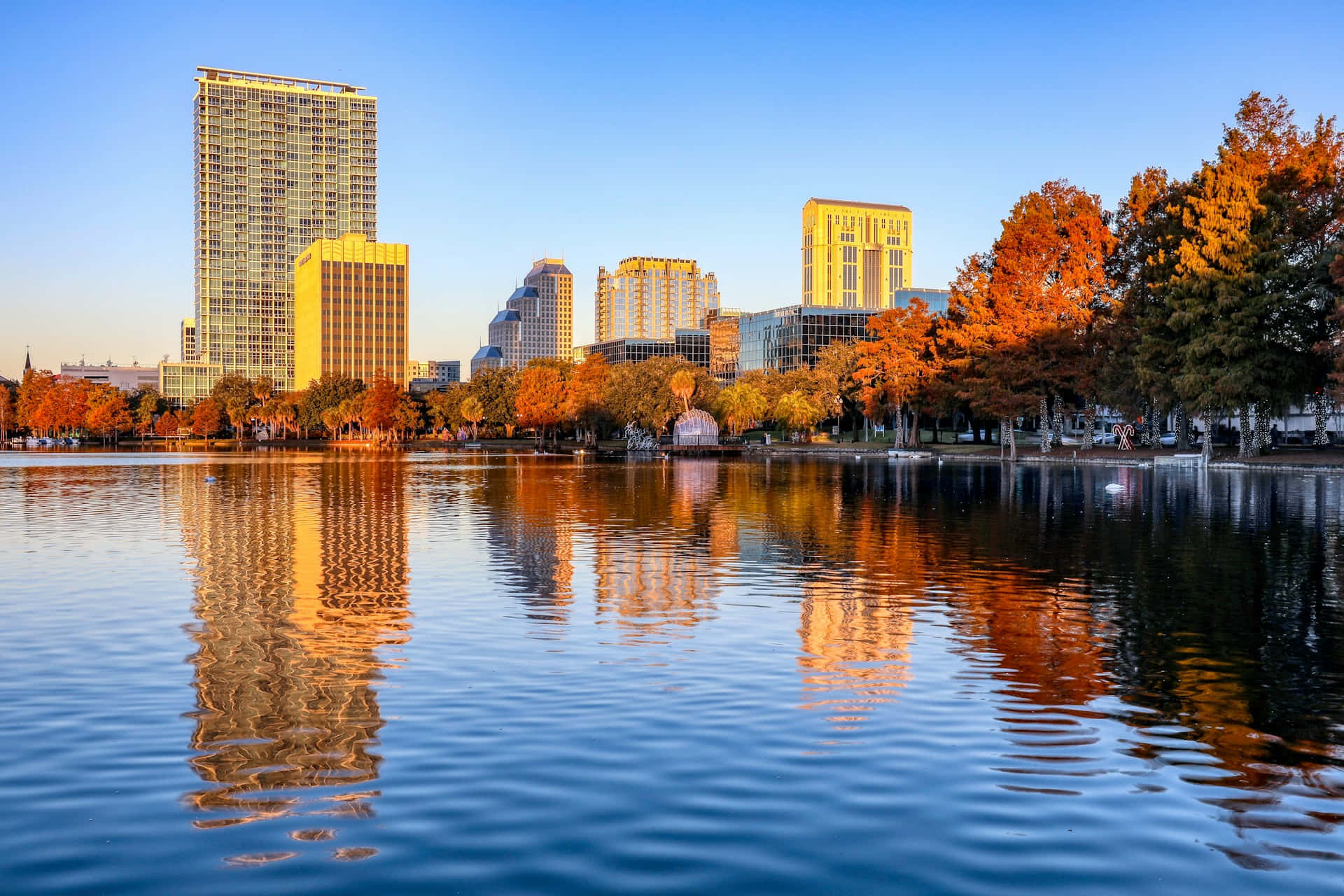 Orlando 桌布 壁紙 — 城市天際線與秋樹倒映在平靜的水面上，金色時刻的清澈藍天下。