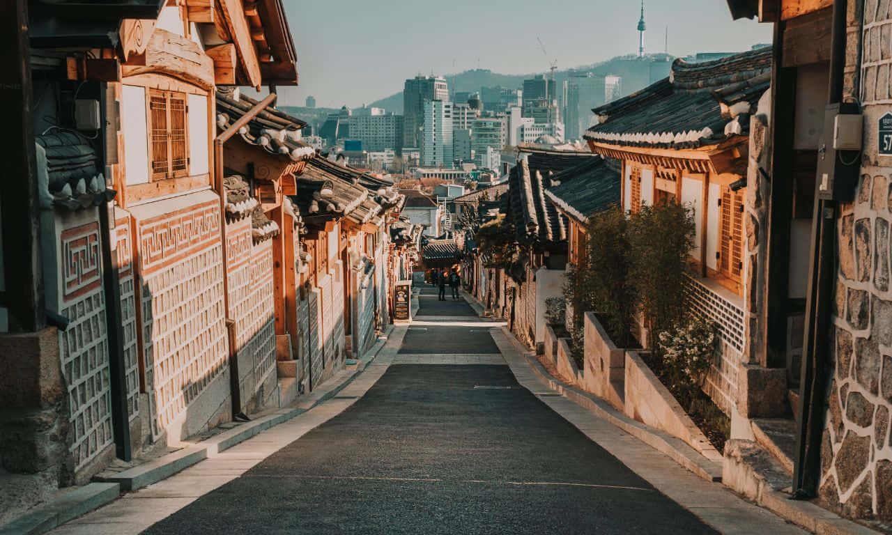 城市燈光在河面的倒影 壁紙 — 一條狹窄的鵝卵石街道，兩旁是傳統的韓國韓屋，通向晴朗藍天下現代化的城市天際線。