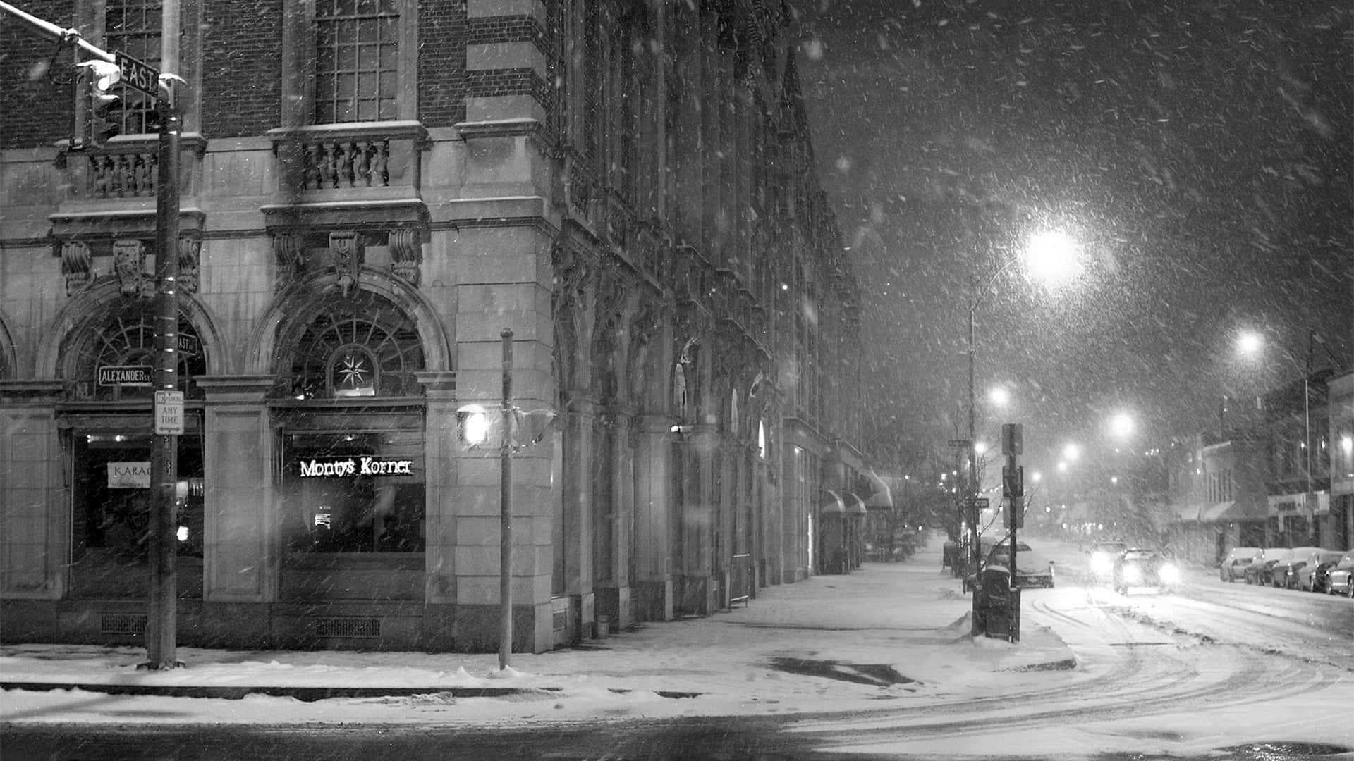 夜間城市街道上飄雪，有明亮的店面和停泊的汽車。