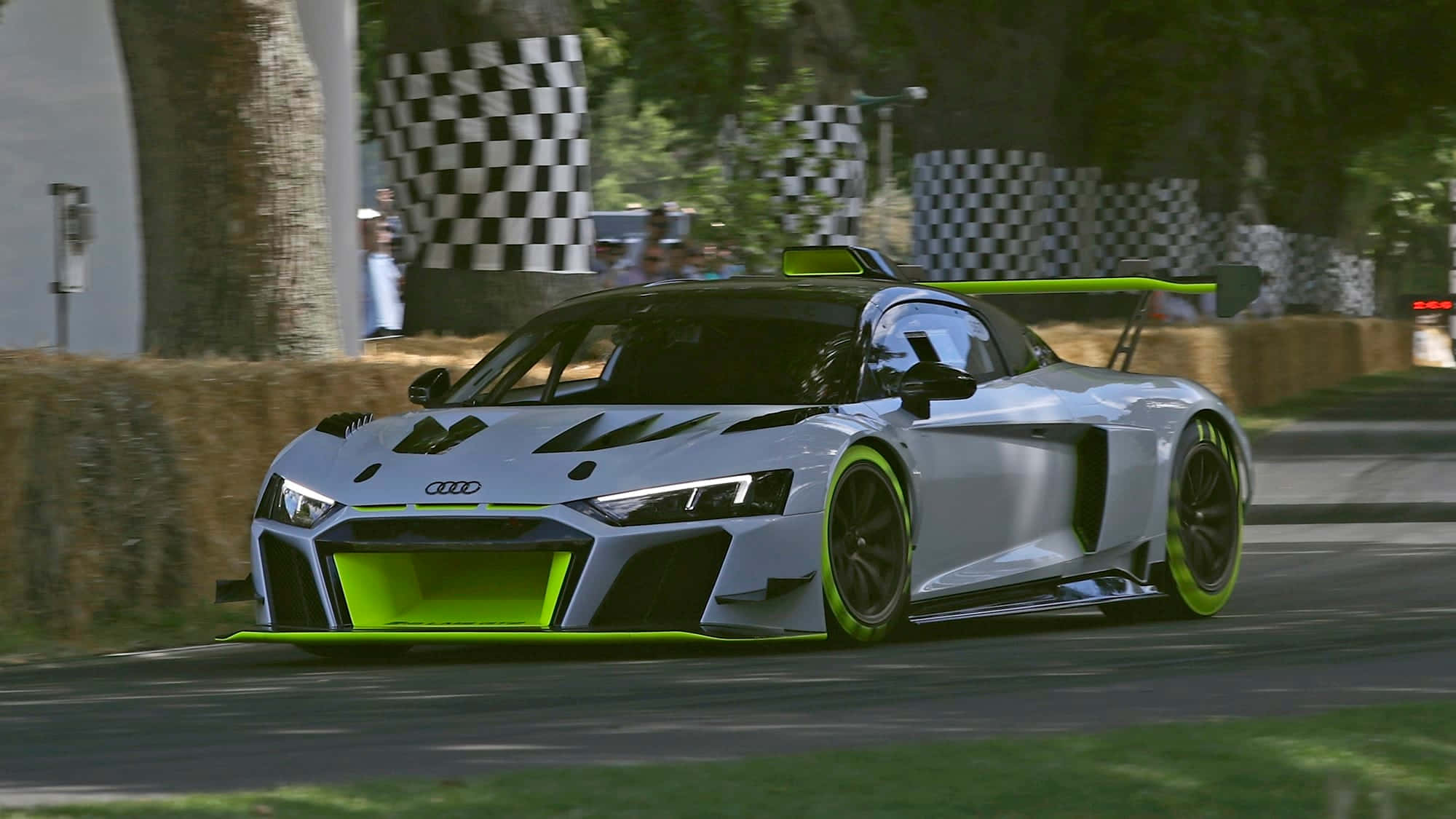 城市駕馭奢華——奧迪A3 壁紙 — 一輛銀色奧迪R8 LMS GT3賽車帶有螢光綠色點綴，在陽光明媚的賽道上飛馳而過乾草捆和方格旗。