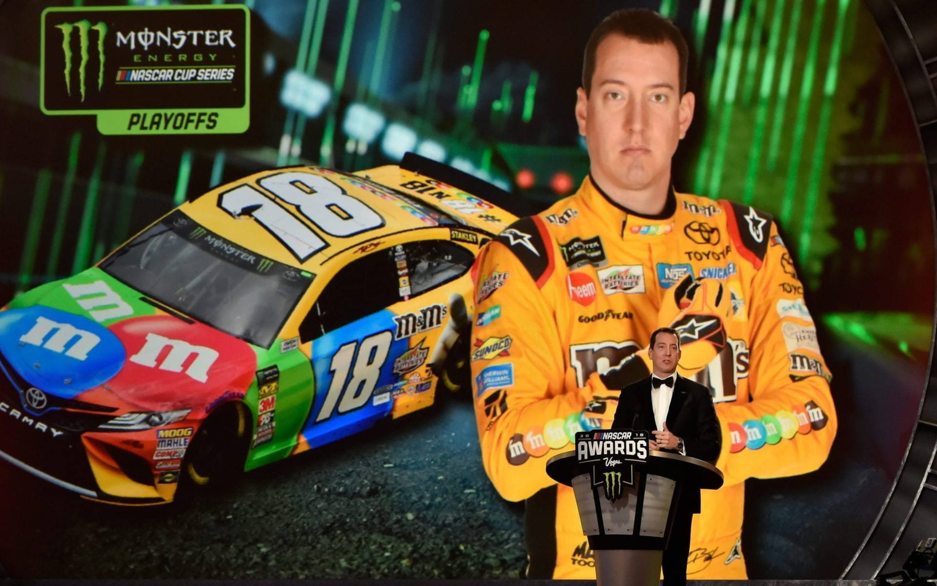 基爾·布希與賽車合影 壁紙 — 一位身穿鮮黃色M&M's賽車服的NASCAR車手，在Monster Energy Awards活動中站在他色彩繽紛的18號賽車旁。