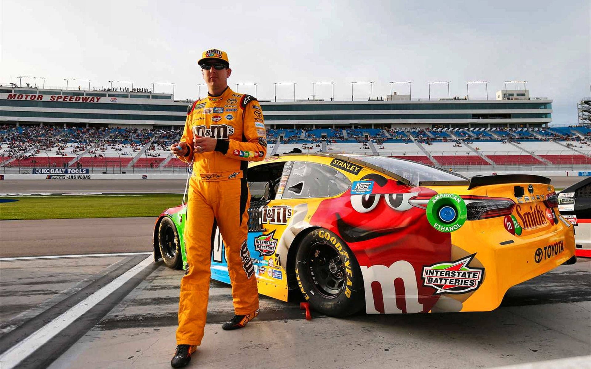 基爾·布希身穿黃色賽服 壁紙 — 一名身穿橙色M&M's賽車服的NASCAR車手站在擁擠的賽車道上，旁邊是他色彩繽紛的M&M's主題Toyota賽車。