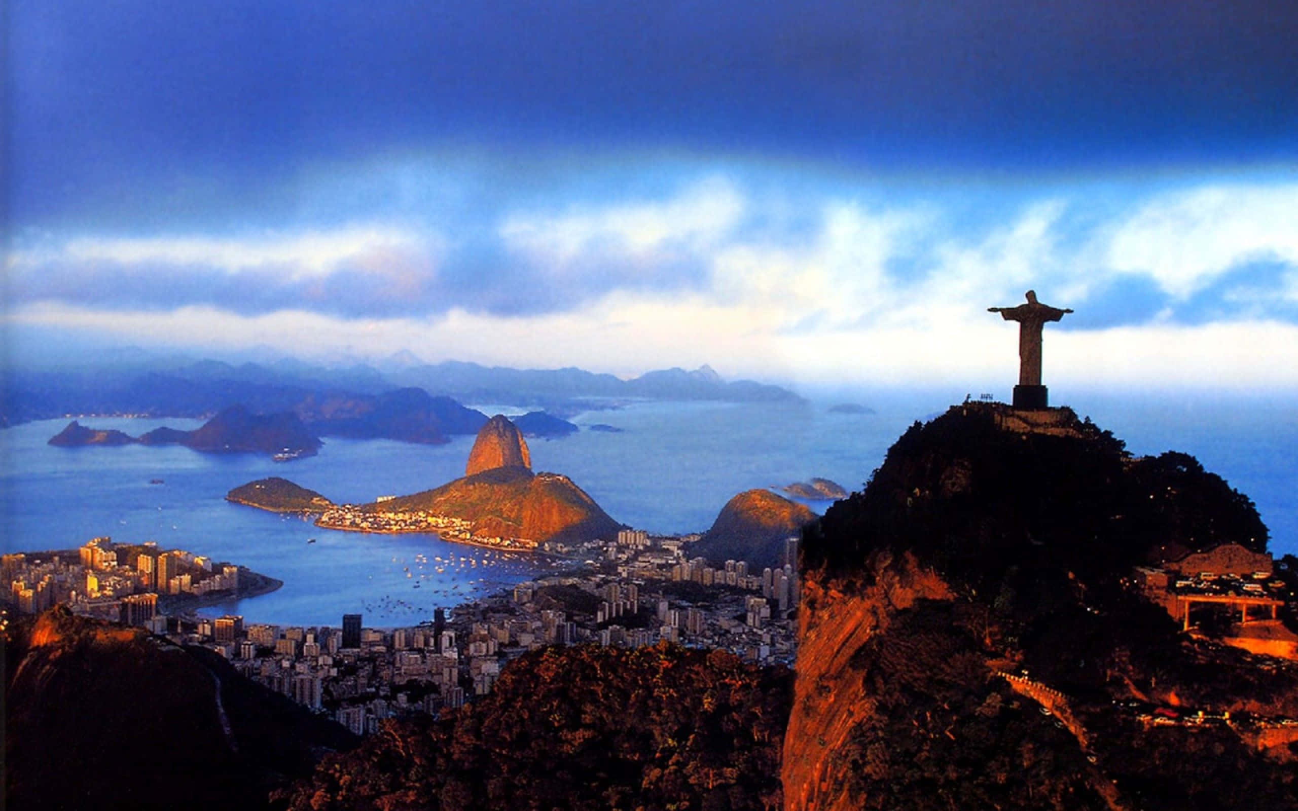 Brazil 桌布 壁紙 — 基督救世主雕像俯瞰日落時分的里約熱內盧，甜麵包山和瓜納巴拉灣沐浴在金色光芒中。