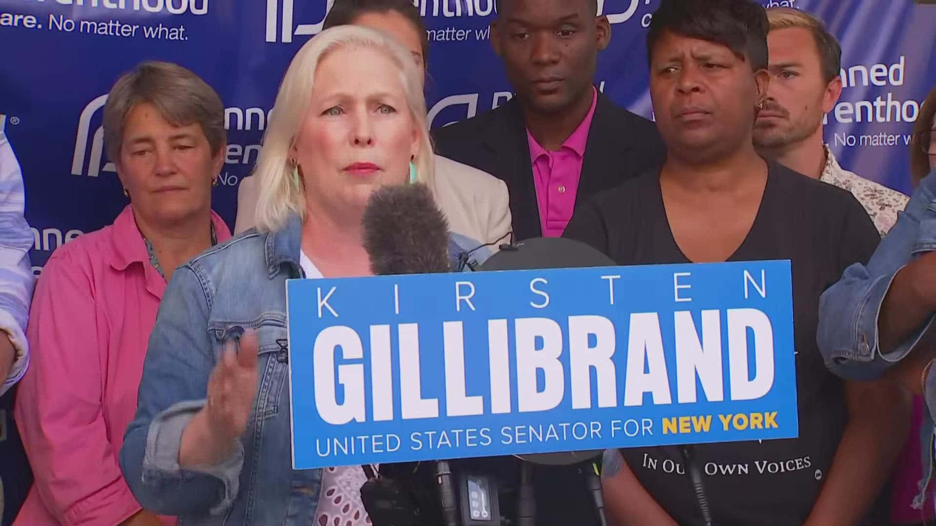 基絲汀·吉爾布蘭德參議員選戰活動 壁紙 — Kirsten Gillibrand 在競選活動中發言，手持藍色標語牌，身後有支持者。