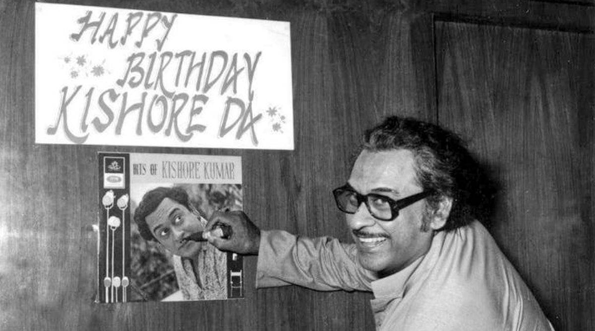 戴眼鏡的男士微笑著指向Kishore Kumar生日海報和唱片。