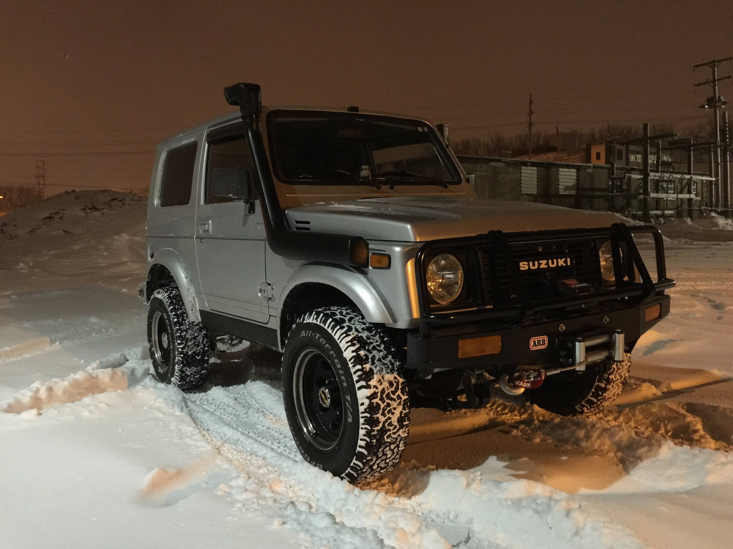 堅固Suzuki 4x4 全地形車 壁紙 — 一輛銀色鈴木Jimny SUV，輪胎被雪覆蓋，停在深雪中，夜空昏暗。