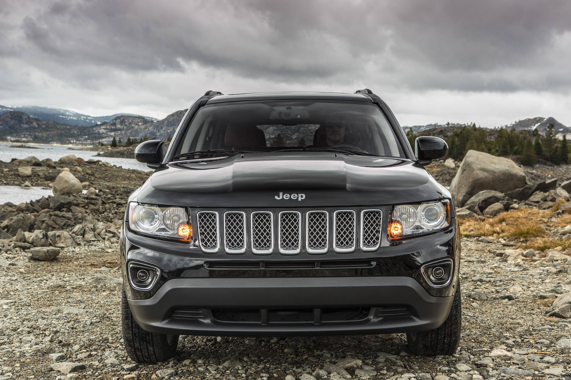 黑色Jeep SUV停在水邊的岩石海岸上，背景有山。