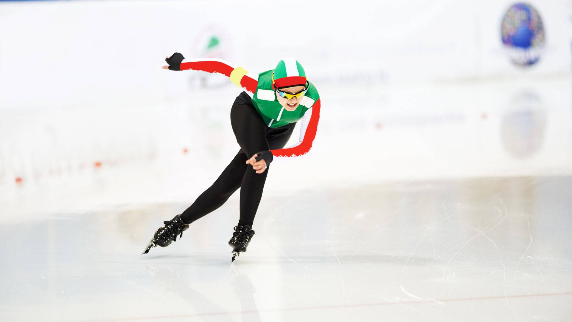堅定的女子速滑選手 壁紙 — 一名穿著綠色、紅色和白色裝備的速度滑冰選手在明亮的冰場上傾身轉彎，背景模糊。