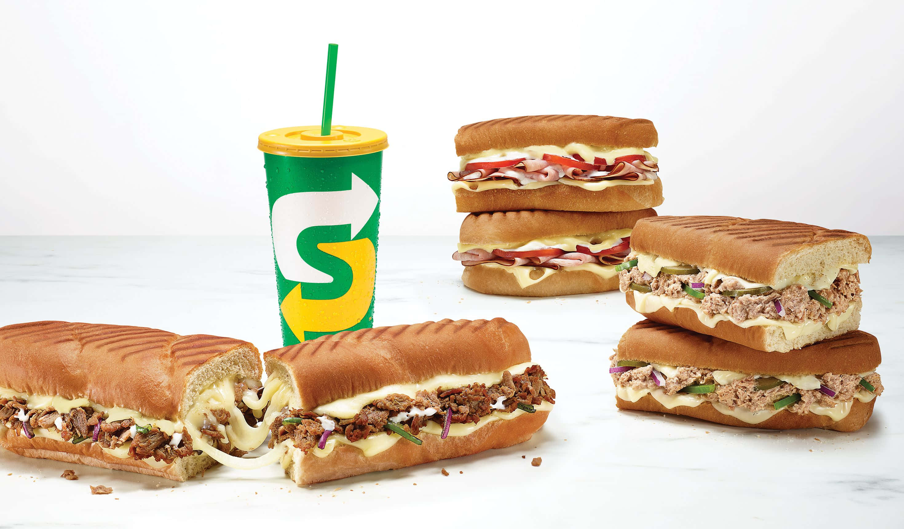 Subway 桌布 壁紙 — 堆疊和切片的三明治，配有肉醬、芝士和蔬菜，旁邊放著一個品牌飲料杯，在乾淨的白色表面上。