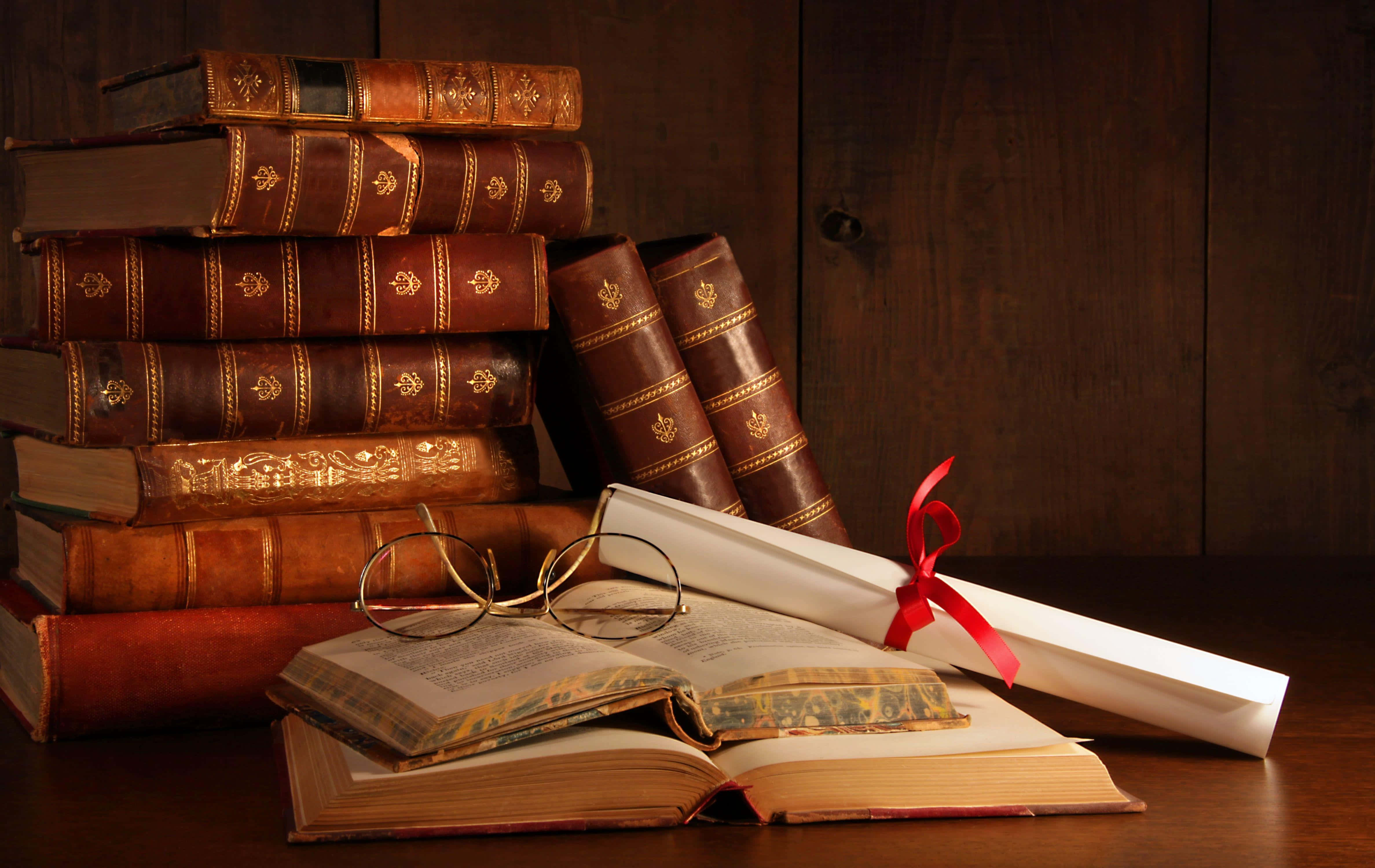 books 桌布 壁紙 — 堆疊的復古皮革裝訂書籍和一本打開的書卷，附有眼鏡和文憑絲帶，放在深色木桌上。