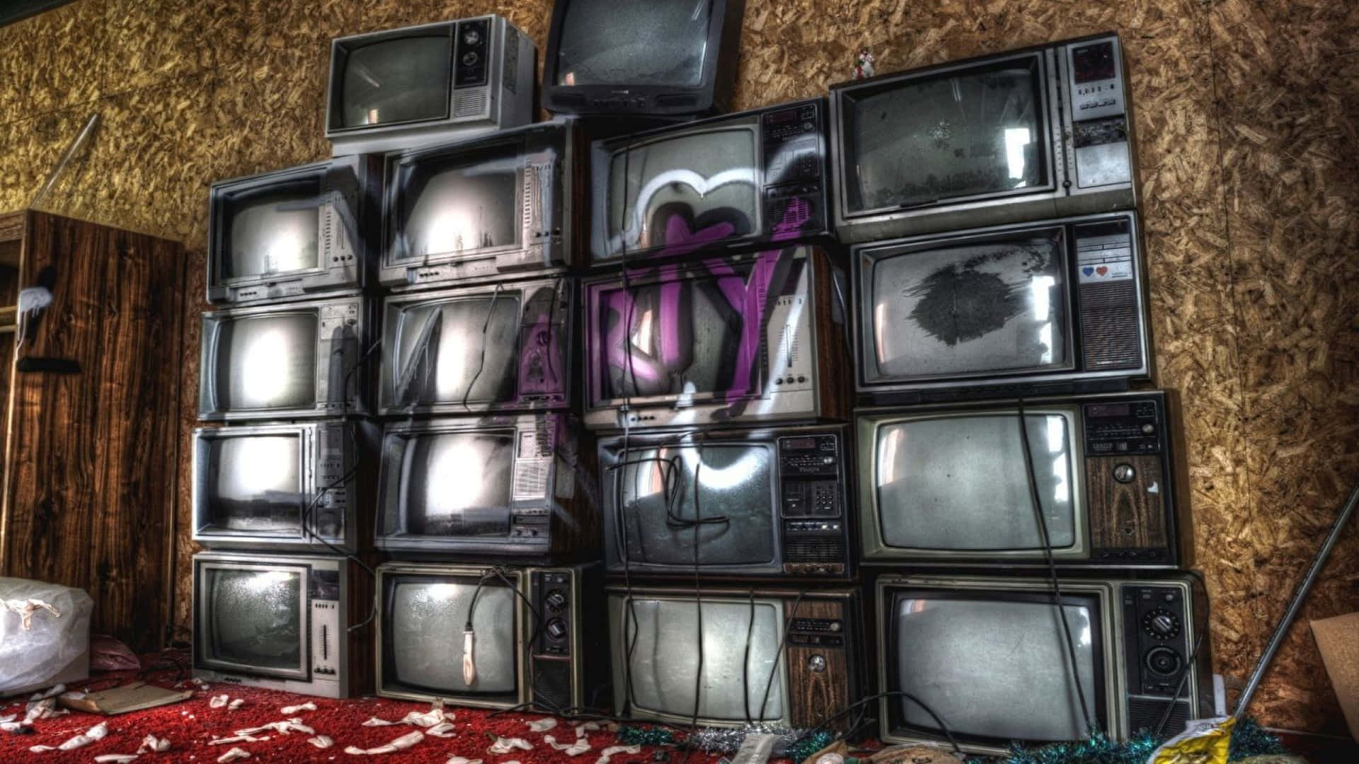 Television Set 桌布 壁紙 — 堆疊的復古電視機，帶有塗鴉和破損屏幕，位於雜亂的房間內，有裸露的OSB牆壁和紅色地毯碎屑。