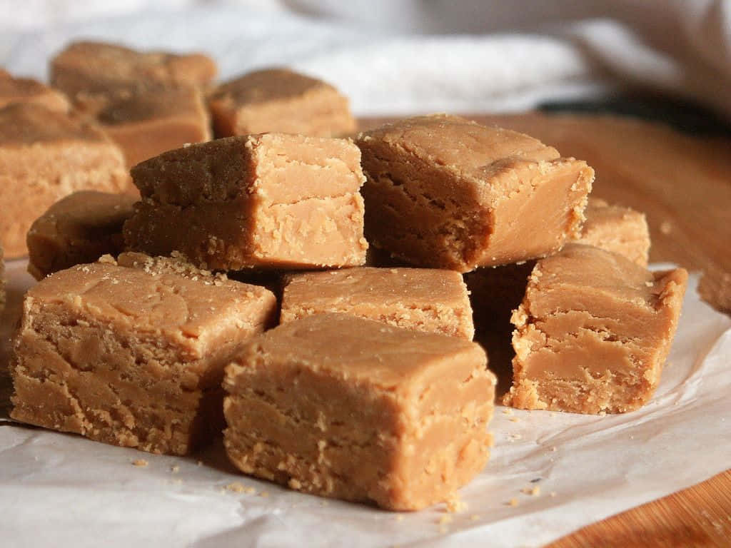Penuche Fudge 桌布 壁紙 — 堆疊的金棕色軟糖方塊，質地易碎，放在木質表面的烘焙紙上。