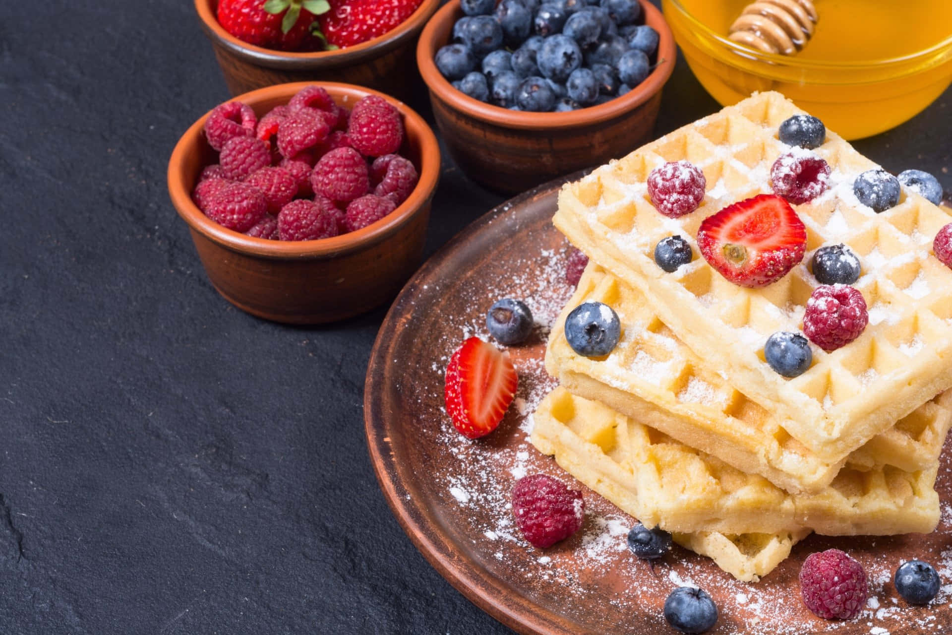 Waffle 桌布 壁紙 — 堆疊的金黃色窩夫撒上糖霜，並放上新鮮漿果，放在鄉村風格的碟子上，旁邊有水果和蜂蜜碗。