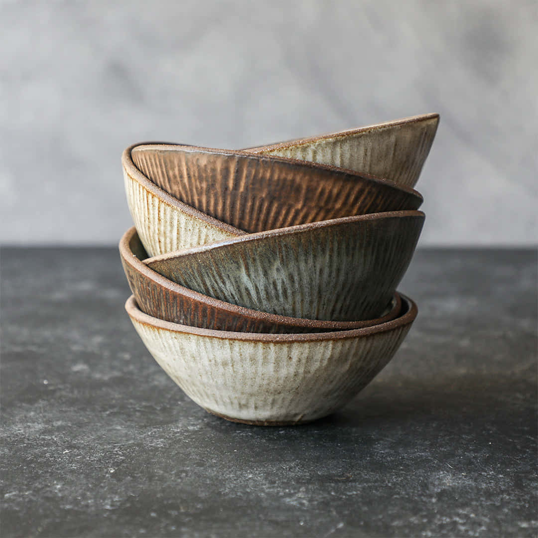 Ceramic 桌布 壁紙 — 堆疊的陶瓷碗帶有紋理脊線和大地色釉料，放置在深色石面上，背景為柔和的灰色。