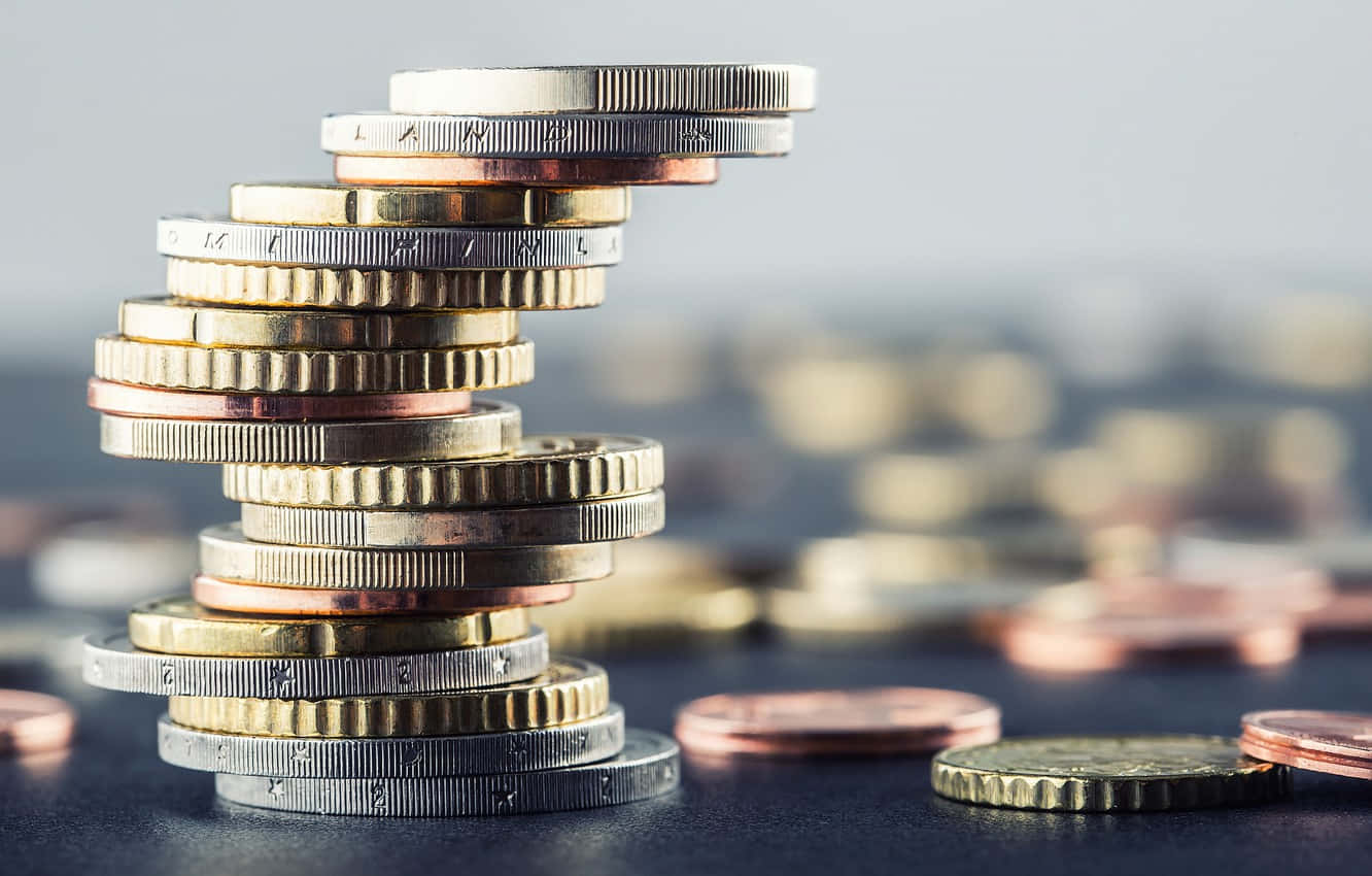 堆疊的黃金、銀幣與銅幣 壁紙 — 一座由歐洲硬幣堆疊而成、搖搖欲墜的螺旋塔，在模糊的散落貨幣背景襯托下聳立。