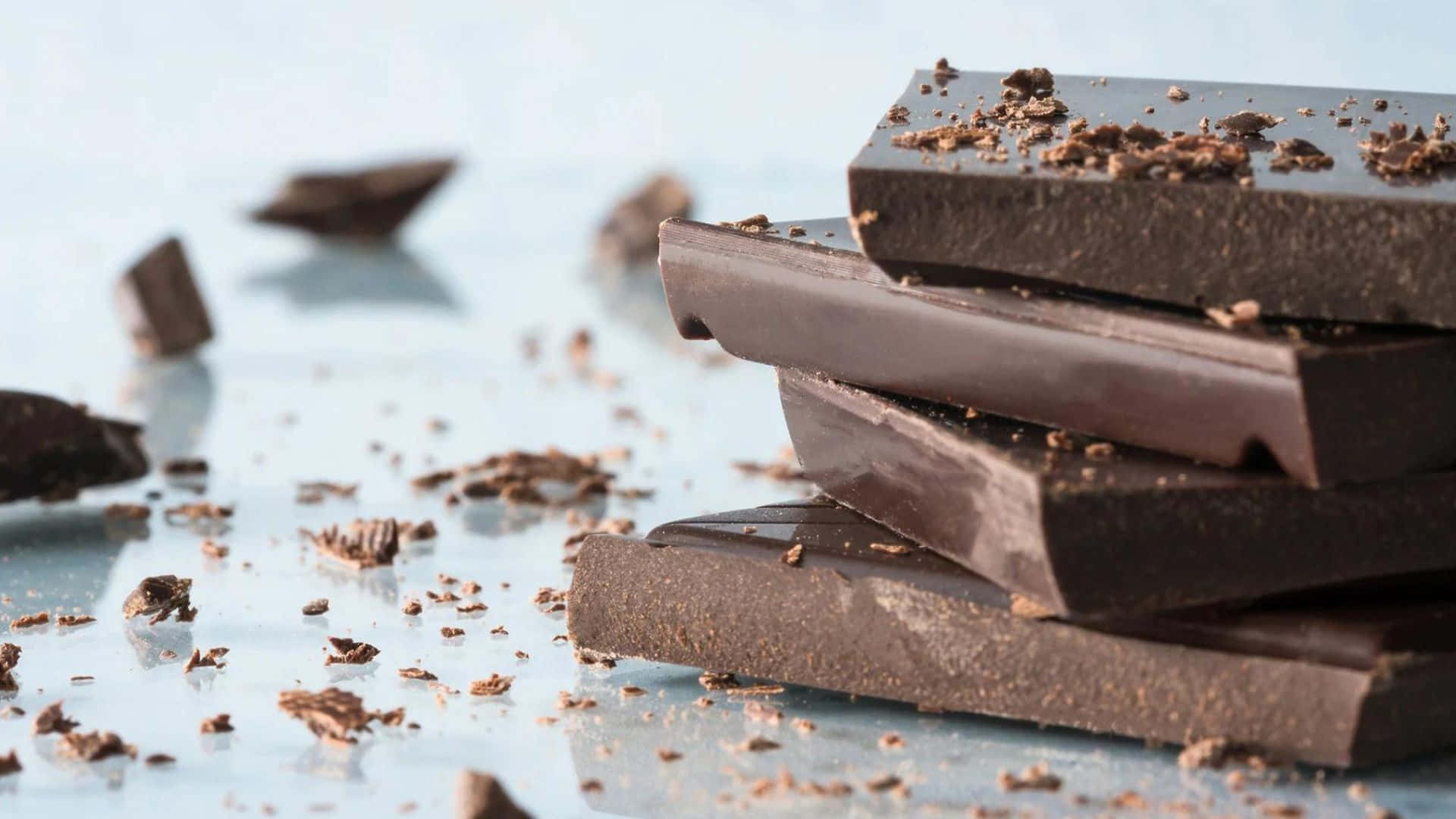 Dark Chocolate 桌布 壁紙 — 堆疊的黑巧克力條，散落著碎屑，放在清涼的藍色反光表面上。