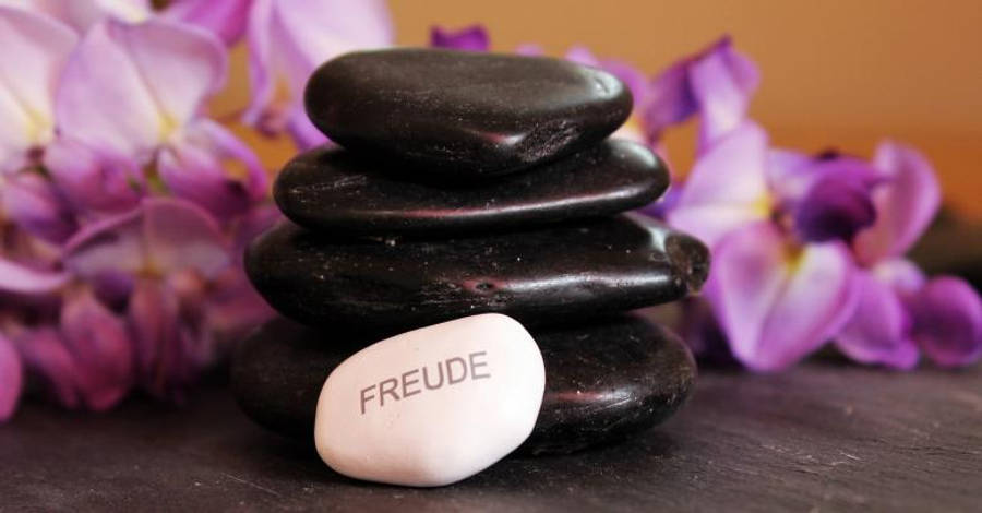堆疊石塔冥想 壁紙 — 一堆光滑的黑色石頭，底部有一塊標有 freude 的白色石頭，周圍環繞著柔和的紫色花朵。