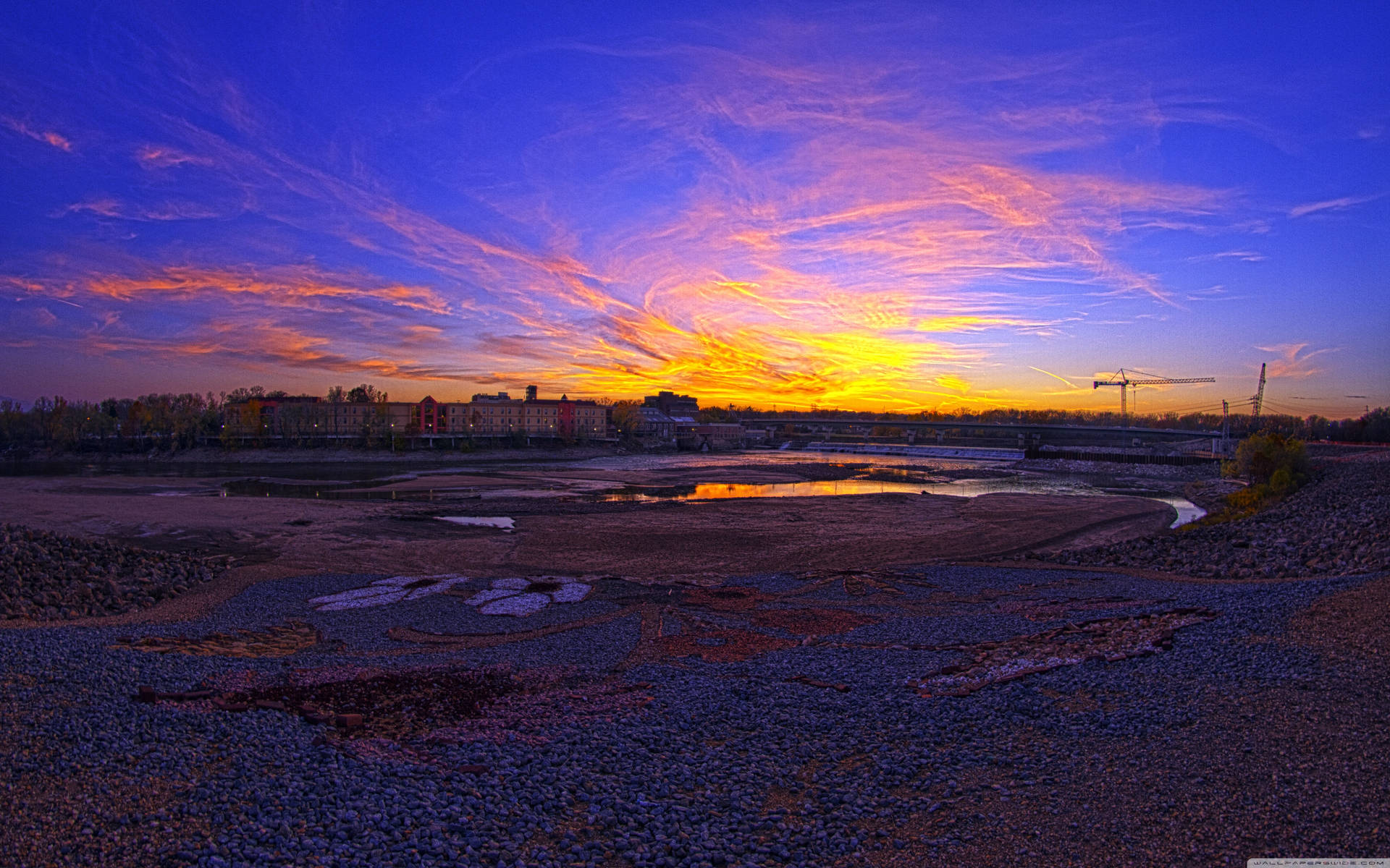 岩石河床、遠處建築物和起重機上方的多彩夕陽。
