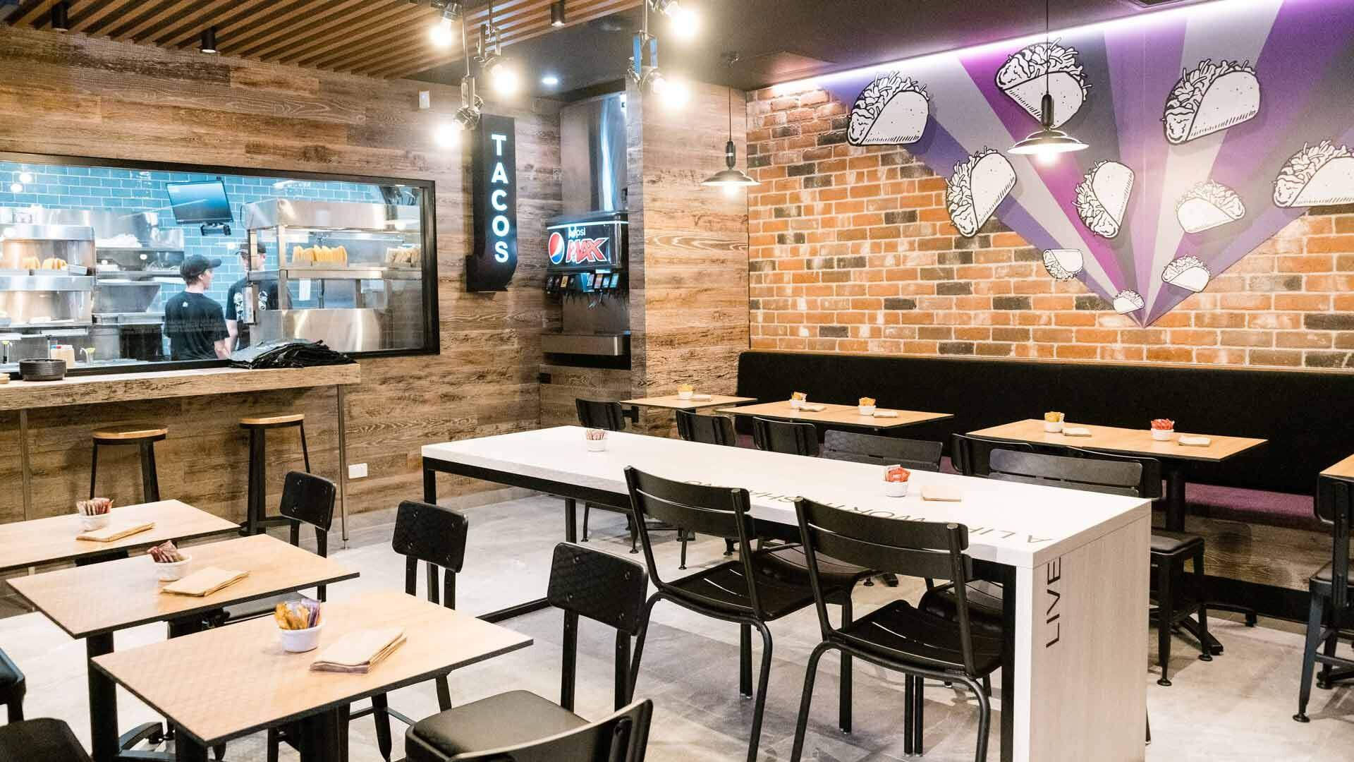 塔可鐘傑斯蒙德分店 壁紙 — 現代墨西哥卷餅餐廳內部，設有磚牆、壁畫、發光燈飾和溫暖吊燈下的木桌。