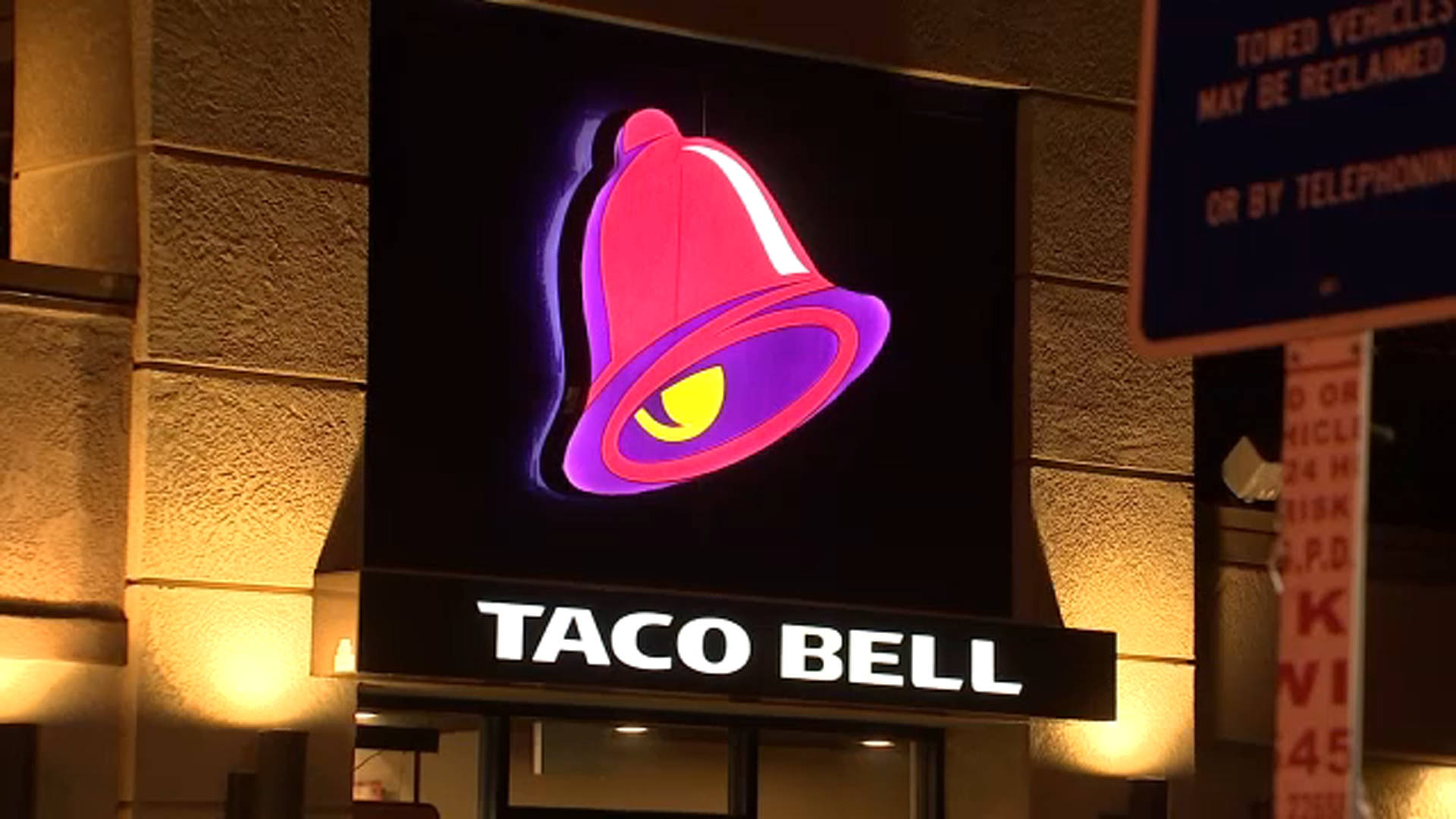 塔可鐘發光招牌 壁紙 — 一個明亮照明的Taco Bell招牌，帶有發光的粉紅色和紫色鈴鐺標誌，在石頭建築外牆上方的黑暗夜空中閃耀。