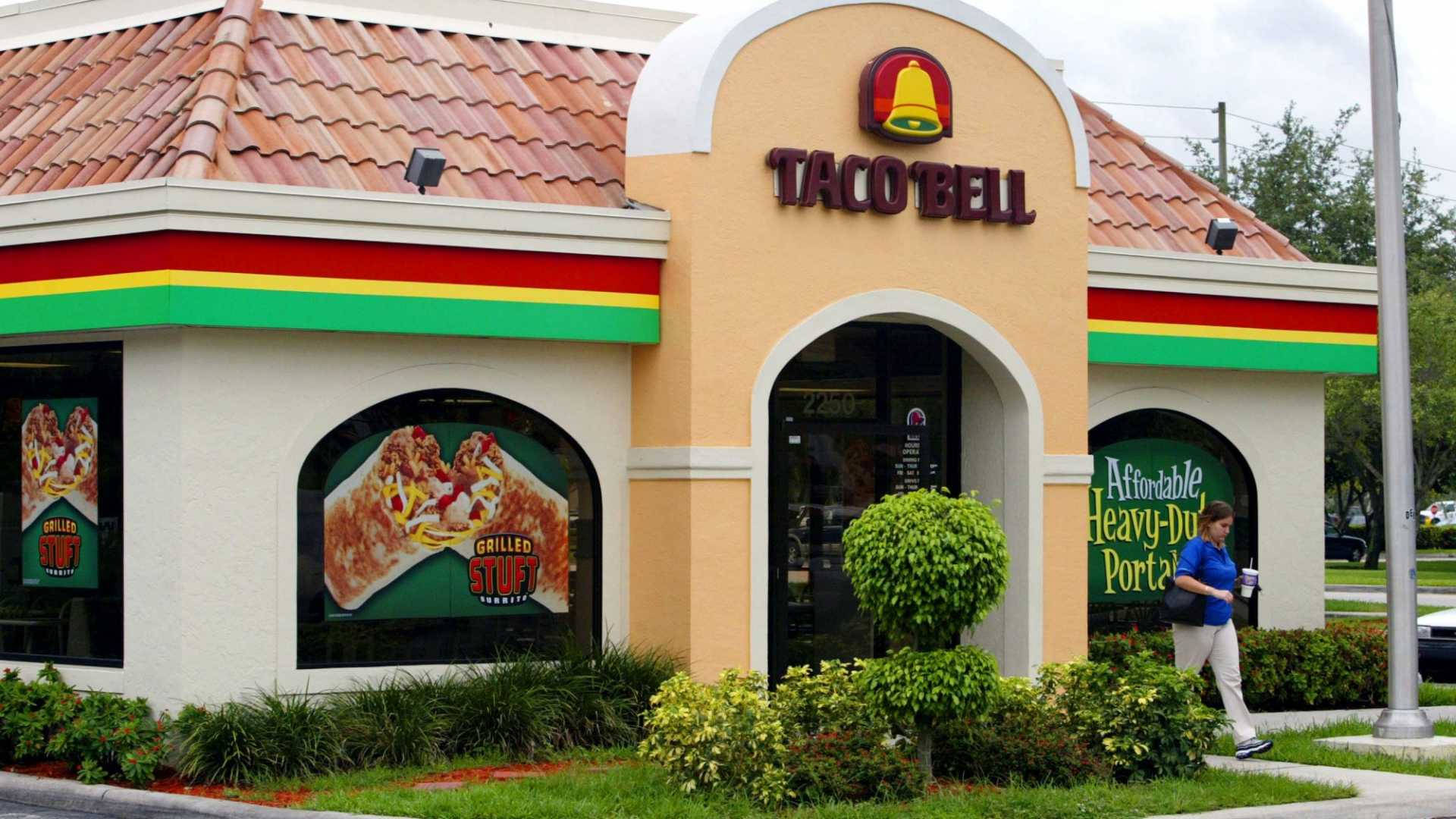 塔可鐘速食餐廳 壁紙 — 一間明亮的Taco Bell餐廳，帶有紅、黃、綠色飾邊，展示大型食物海報，並有一個人行人在人行道上走過。