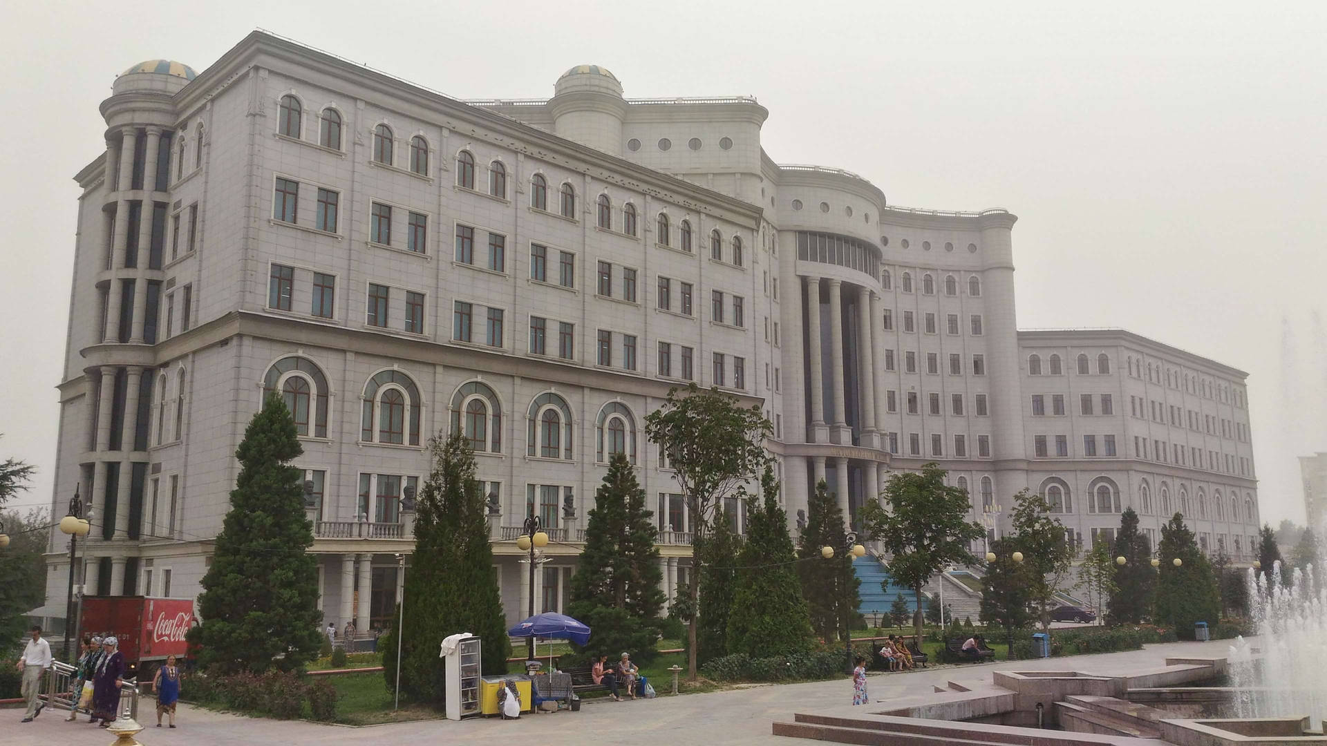 一座帶有拱形窗戶和圓頂的大型白色建築，周圍環繞著樹木和噴泉，廣場上有人們。