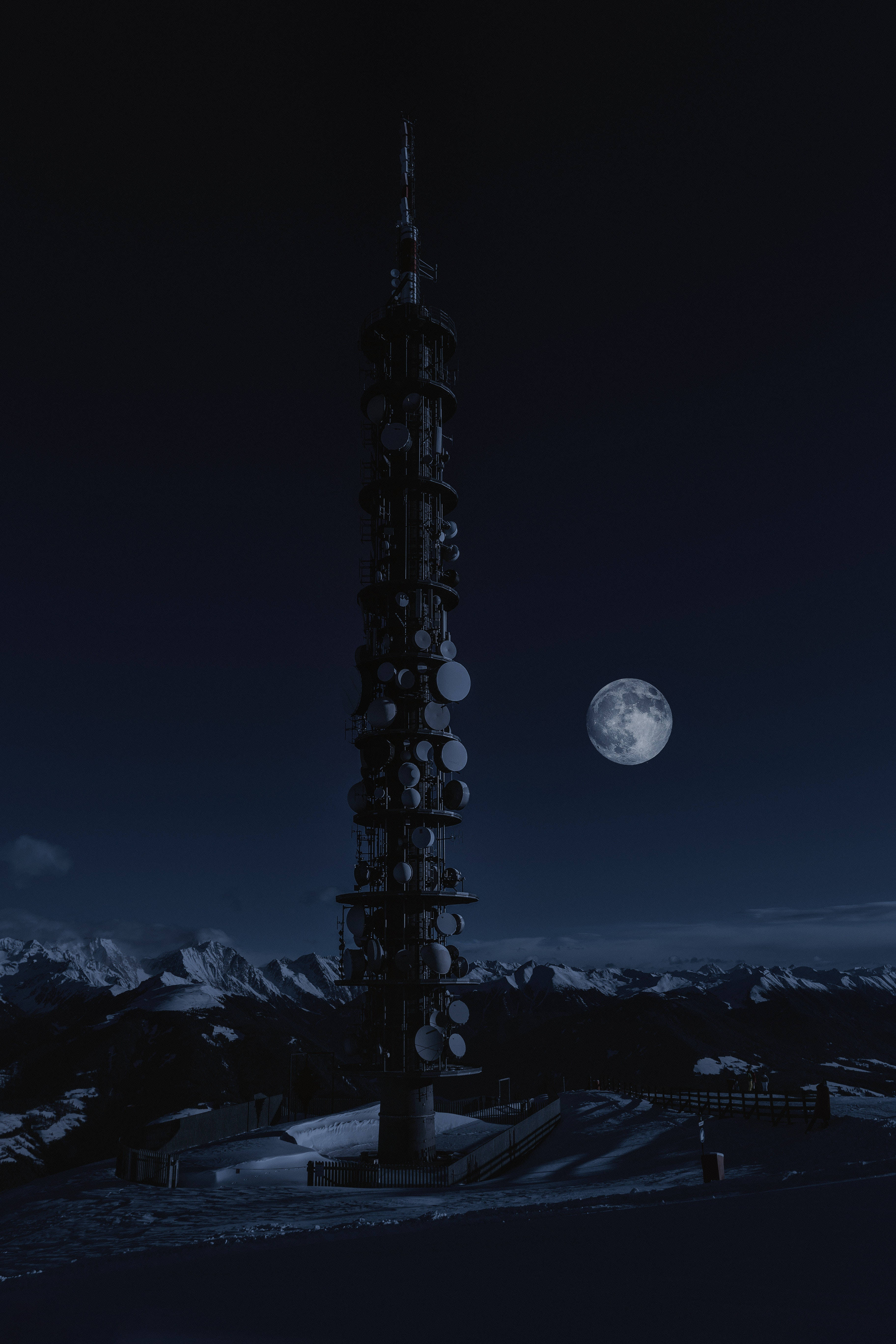 塔旁滿月 壁紙 — 一座高聳的通訊塔在夜間滿月下獨自矗立在積雪的山頂上。