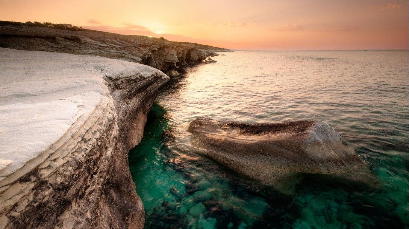 塞浦路斯阿爾馬努海灘 壁紙 — 夕陽將崎嶇的海岸懸崖沐浴在溫暖的金色中，而碧綠的海水輕拍著風化的岩石結構。