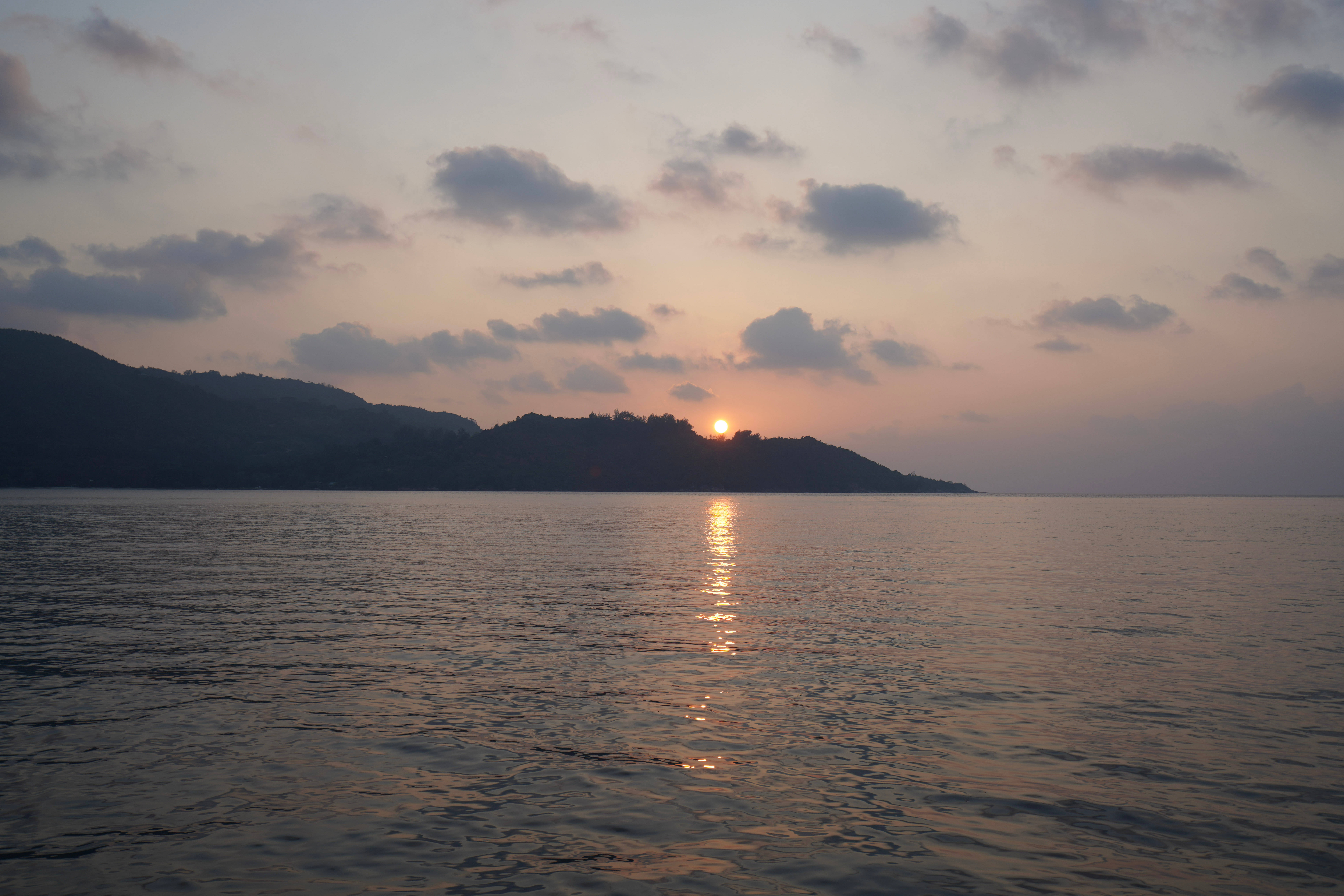 塞舌爾海洋日落 壁紙 — 夕陽溫暖地照耀在平靜的水面上，遠山在柔和的粉紅色和橙色雲彩中形成剪影。