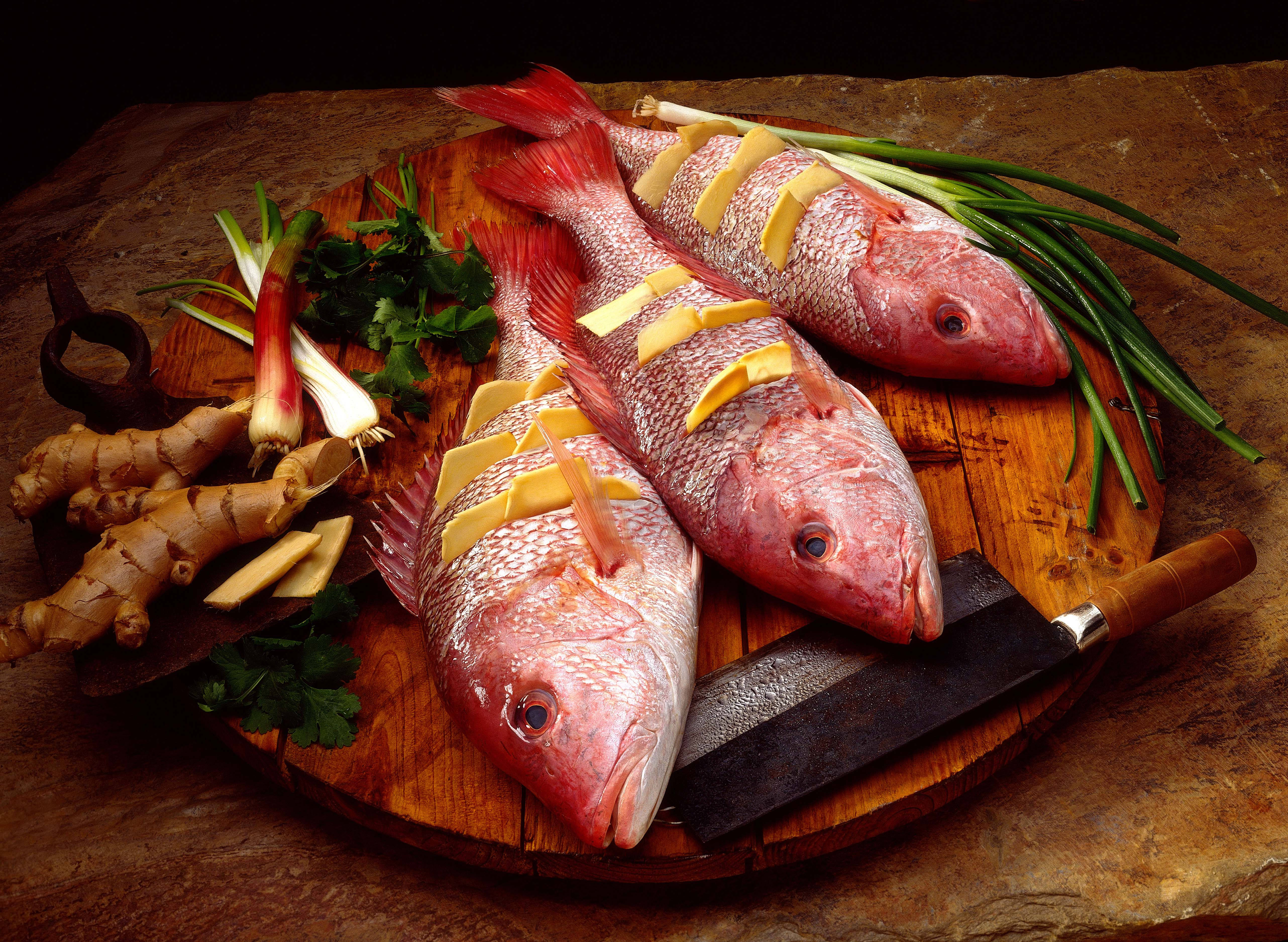 填餡紅鯛魚料理 壁紙 — 三條完整嘅紅鯛魚，用薑同蔥裝飾，放喺一個鄉村風格嘅木砧板上，旁邊有把菜刀。
