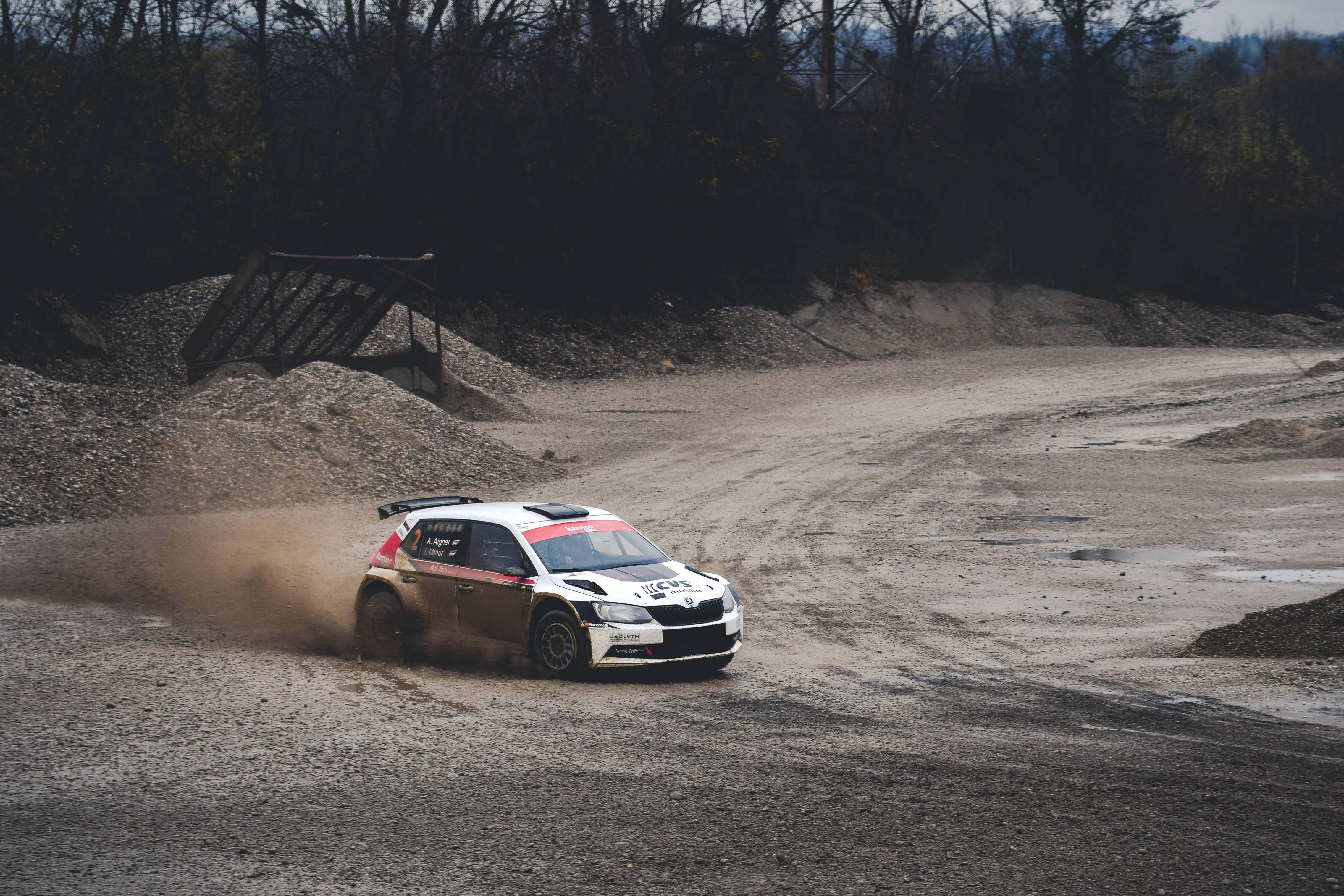 《塵土拉力賽》Skoda R5 競賽畫面 壁紙 — 一輛白色拉力賽車在泥濘的碎石路上疾馳，揚起灰塵，周圍是黑暗的樹木和土堆。