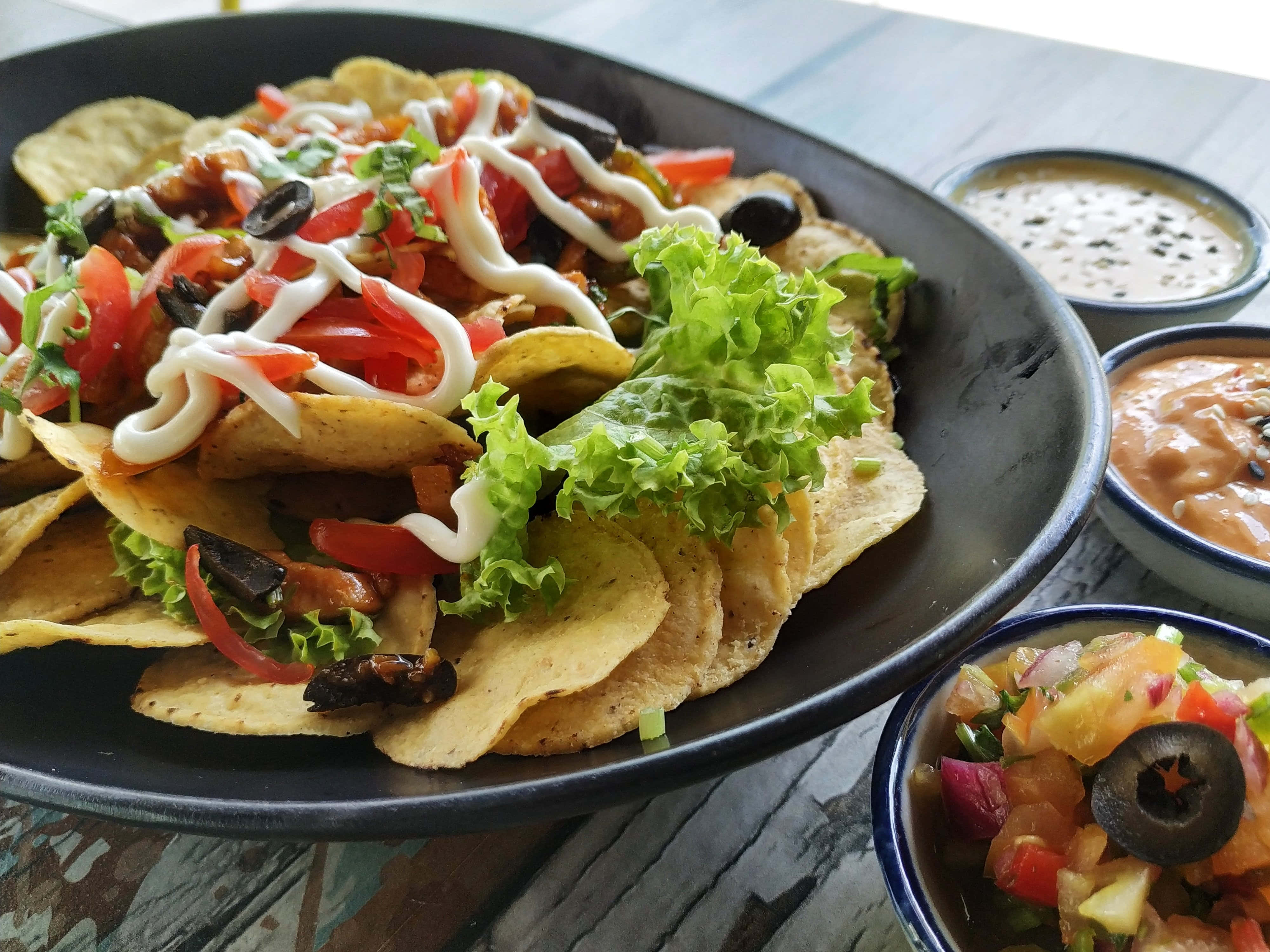 墨西哥 Nachos 食物 壁紙 — 一盤色彩繽紛的滿載玉米片，上面鋪滿新鮮蔬菜、酸奶油和生菜，旁邊有三款蘸醬，放在質樸的木桌上。