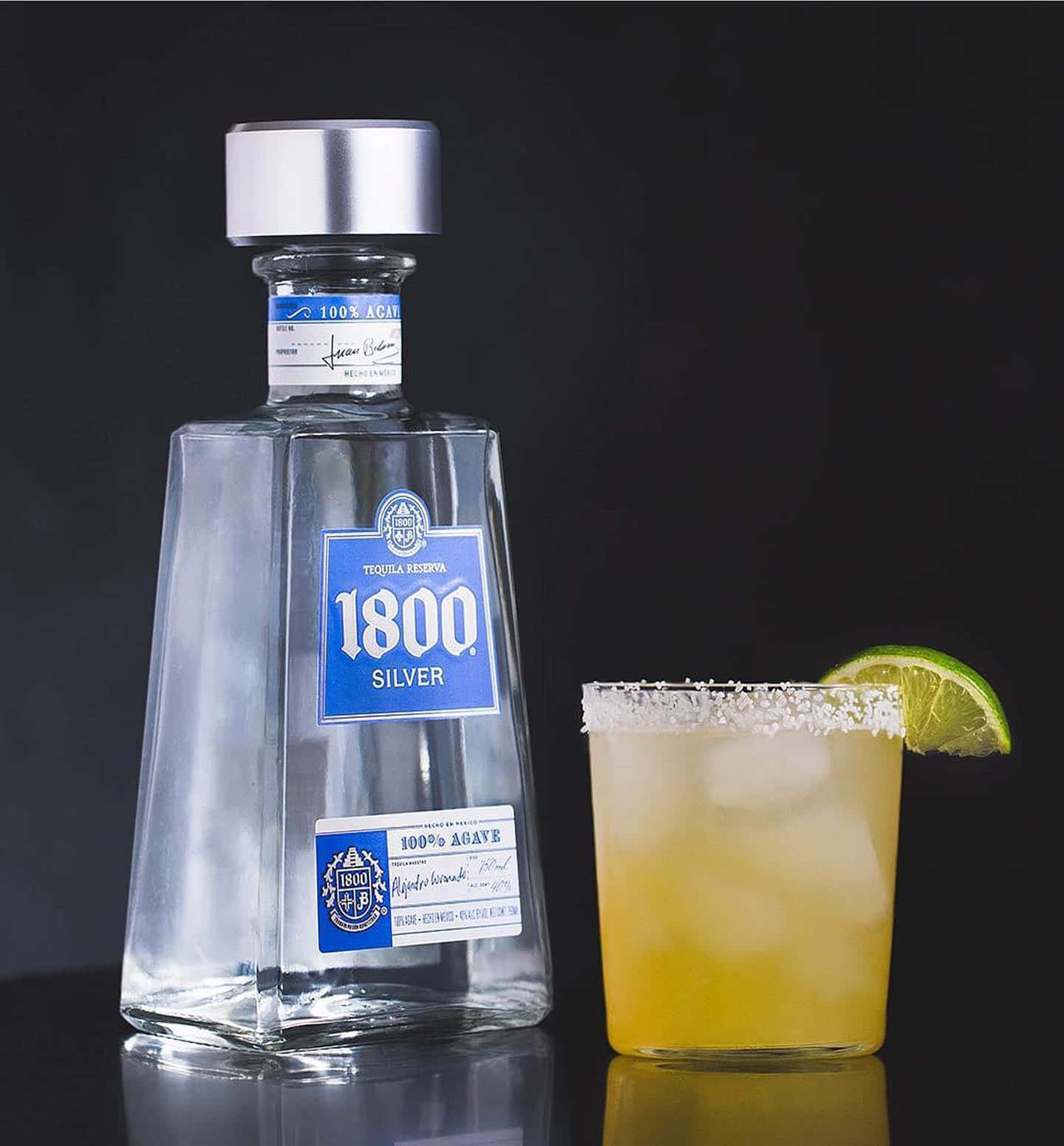 墨西哥品牌1800龍舌蘭酒Reserva Silver與柑橘瑪格麗特雞尾酒 壁紙 — 一個時尚的1800 Silver龍舌蘭酒瓶旁邊放著一杯鹽邊瑪格麗特，配以青檸裝飾，置於深色反光表面上。