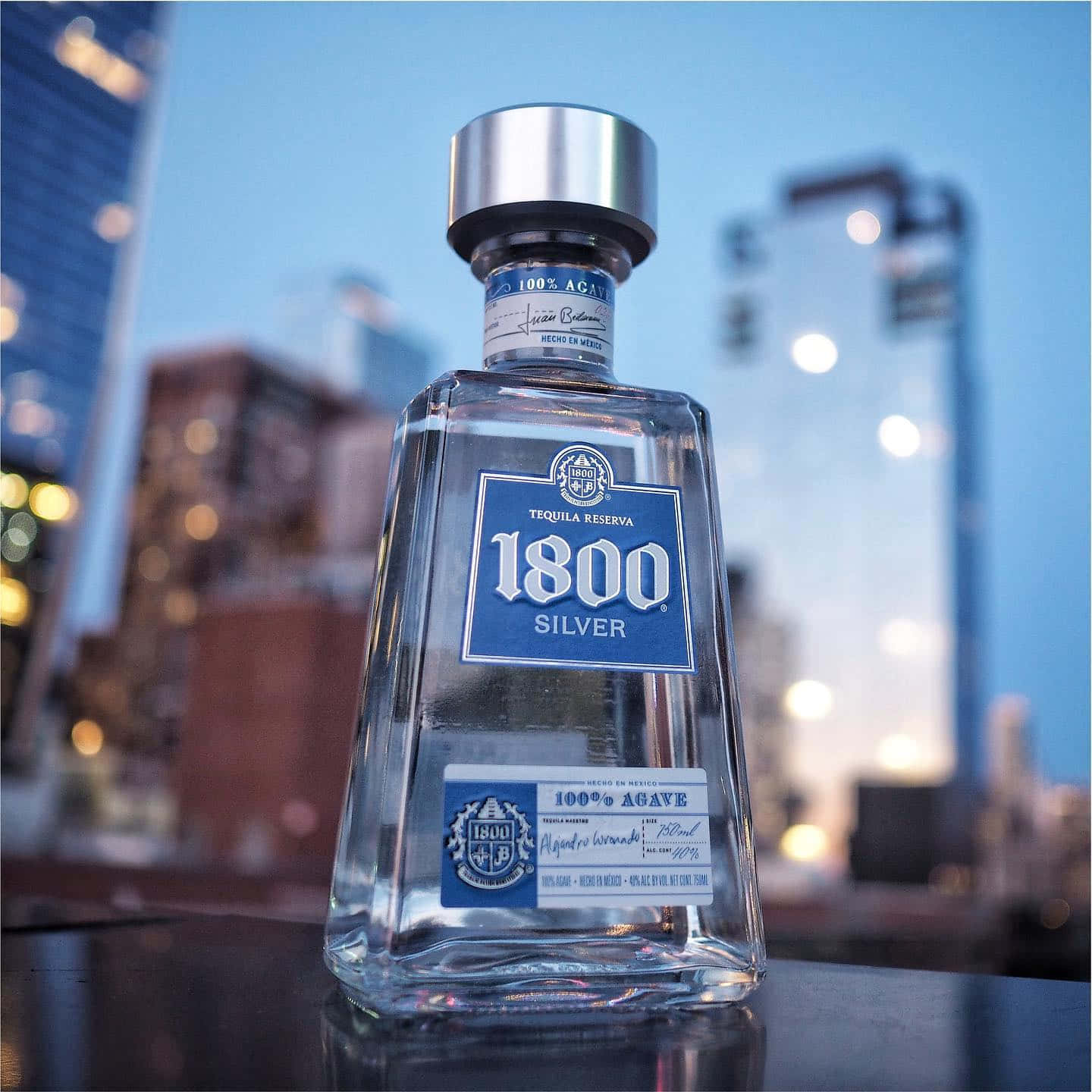 墨西哥品牌1800龍舌蘭酒Reserva Silver城市建築背景 壁紙 — 一個清澈的1800 Silver龍舌蘭酒瓶在屋頂邊緣閃耀，背景是暮色中柔和模糊的城市天際線。
