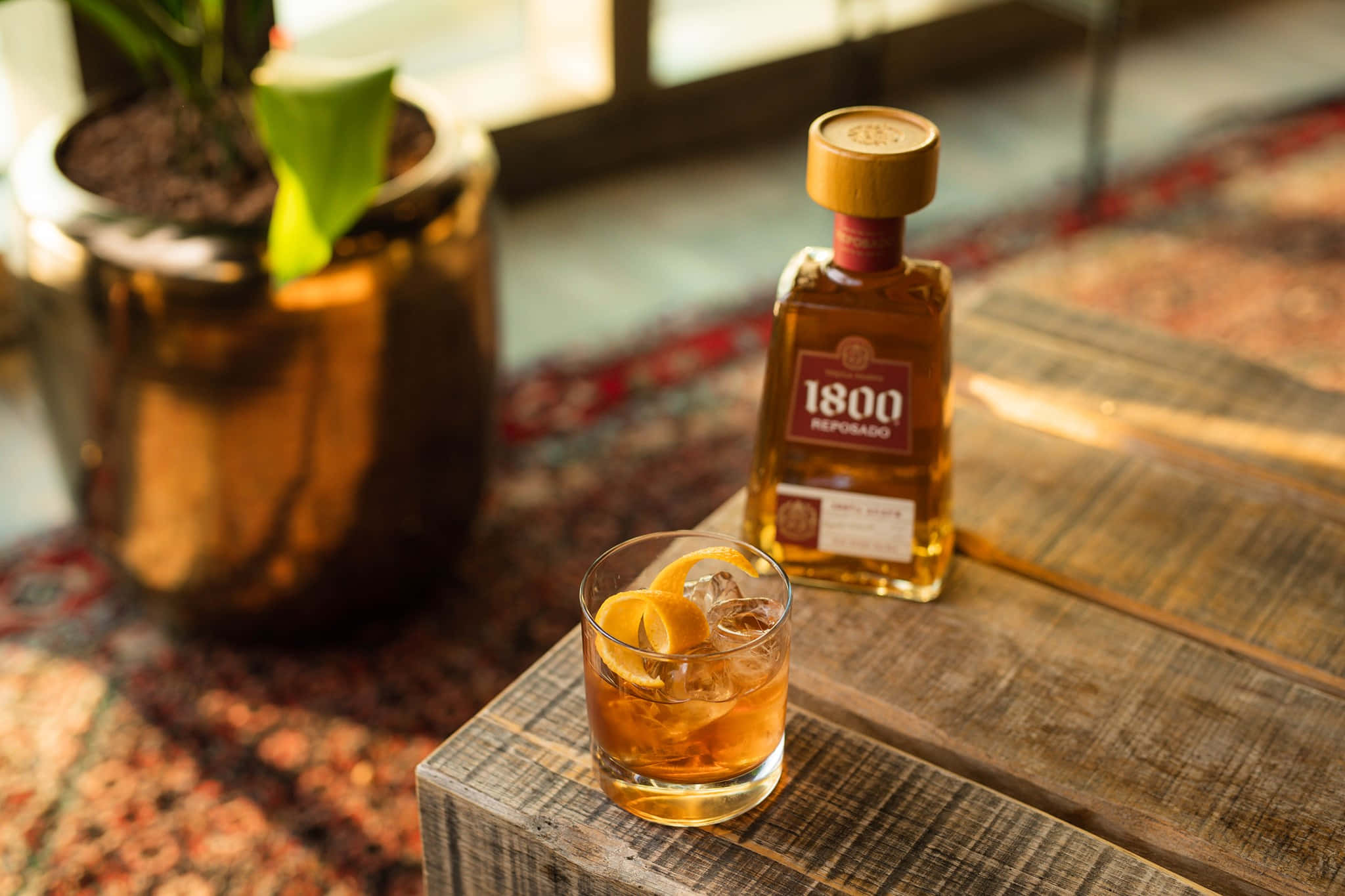 墨西哥品牌1800龍舌蘭酒Reserva Reposado與經典老式雞尾酒 壁紙 — 一杯琥珀色威士忌加冰塊和柑橘裝飾，旁邊放著酒瓶，放在盆栽植物附近的鄉村風格木桌上。