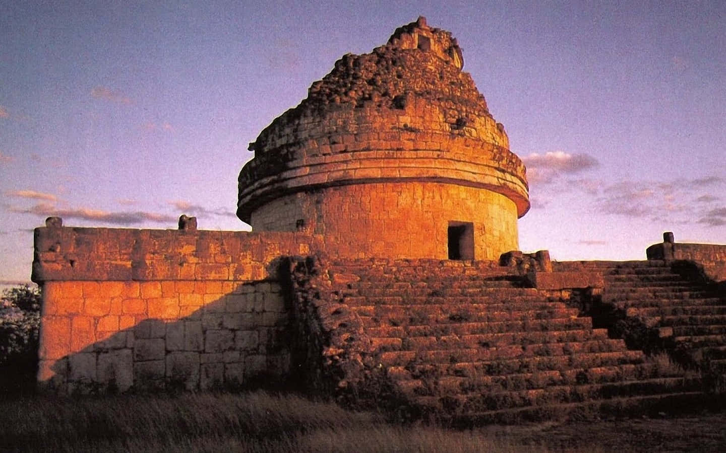 墨西哥奇琴伊察「鷹巢」日落 壁紙 — 古老石廟沐浴在金色夕陽下，長長的影子延伸在風化的台階和草地上。