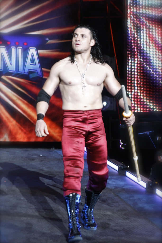 墨西哥摔角手「El Zorro」步入 Wrestlemania 壁紙 — 一位肌肉發達的摔角手，身穿紅色緞面褲子和藍色靴子，手持權杖自信地走過燈光明亮的舞台。